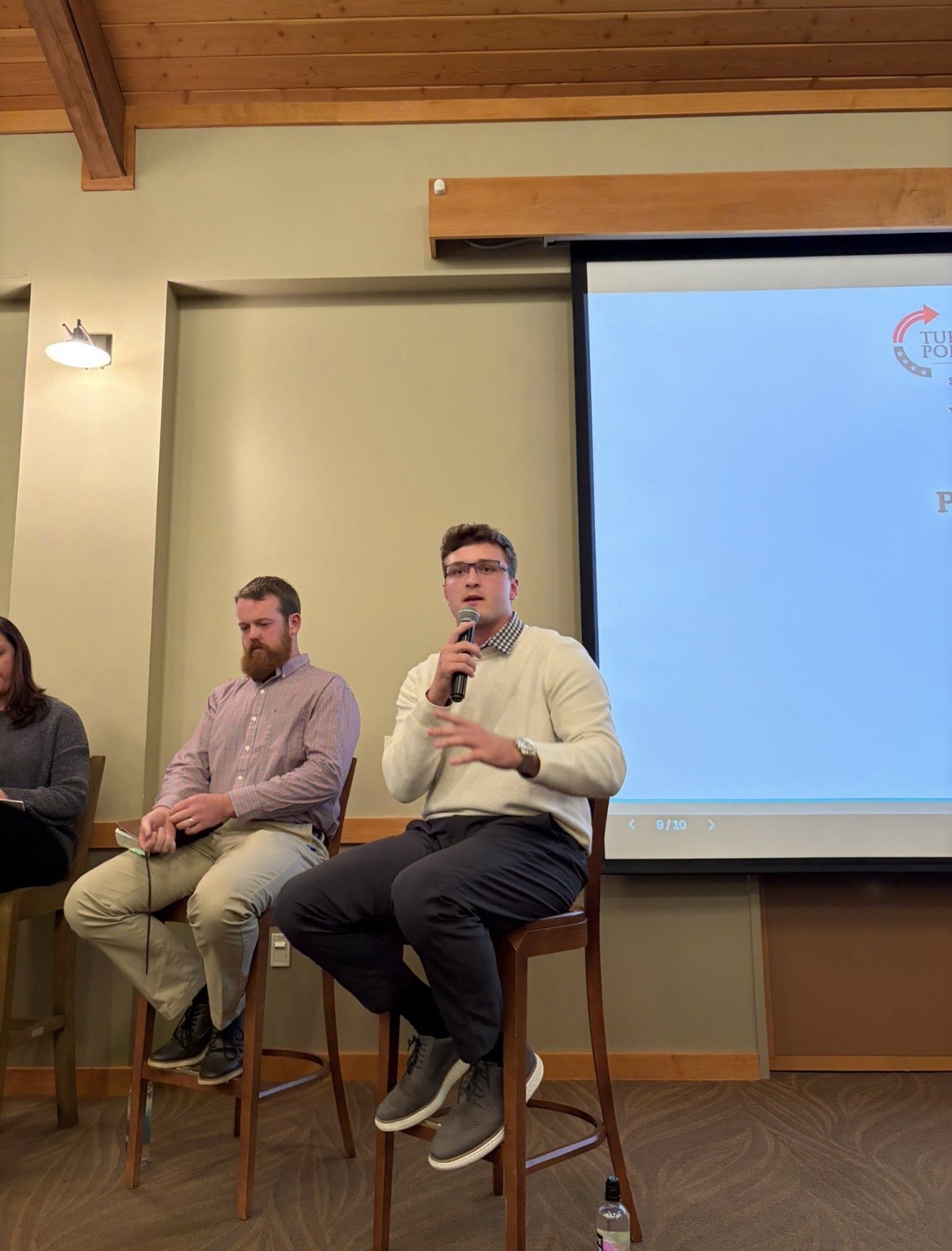 A young man with glasses and a white sweater holding a microphone, sitting on a high stool during a panel discussion, with a large screen displaying a logo in the background.