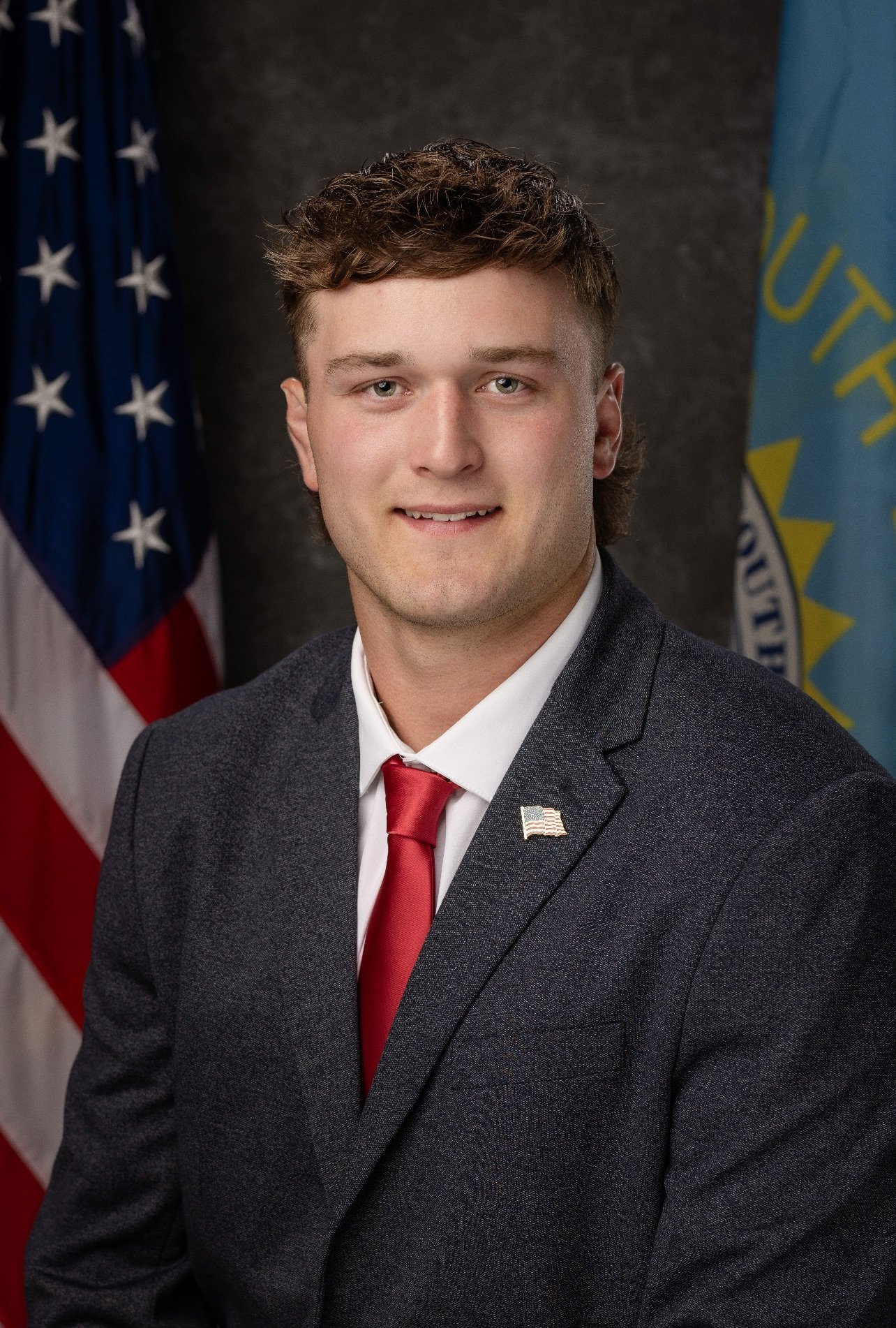 A young man in a gray suit, white shirt, and red tie, wearing a small American flag lapel pin, posing for a formal portrait with the US flag and government emblem flags in the background.