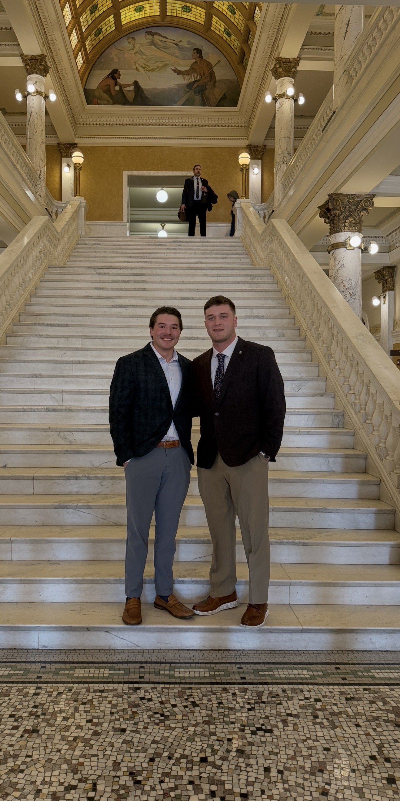 Two young men in blazers and dress shoes standing on a grand marble staircase inside an elegant building with ornate columns and a mural above the staircase, with a man on the landing above taking a photo.