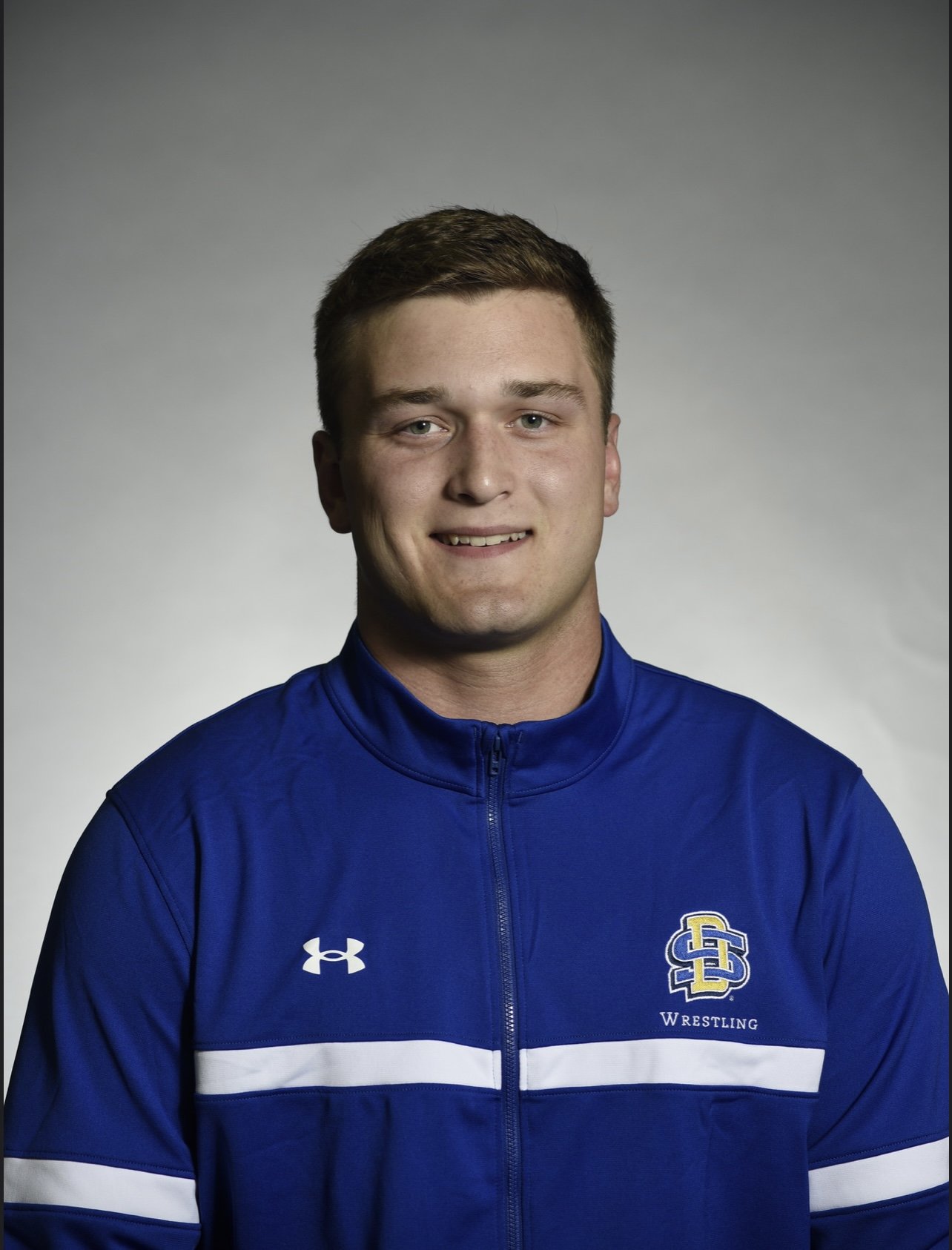 A young man with short brown hair wearing a blue sports jacket with white stripes, an Under Armour logo, and a logo for SD Wrestling on the chest, smiling at the camera.