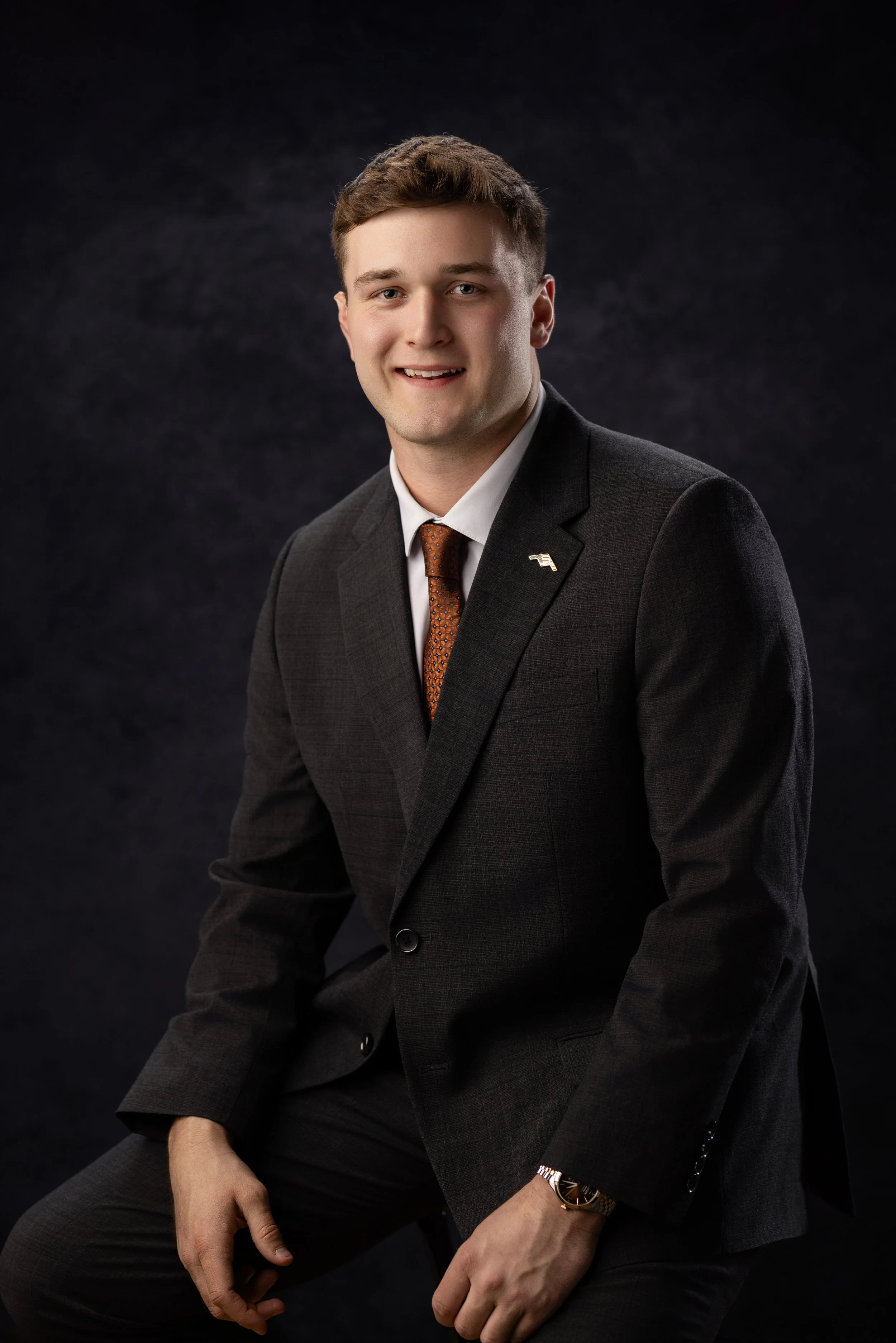 A young man in a dark suit with a white shirt and burnt orange tie, smiling, sitting against a dark background.