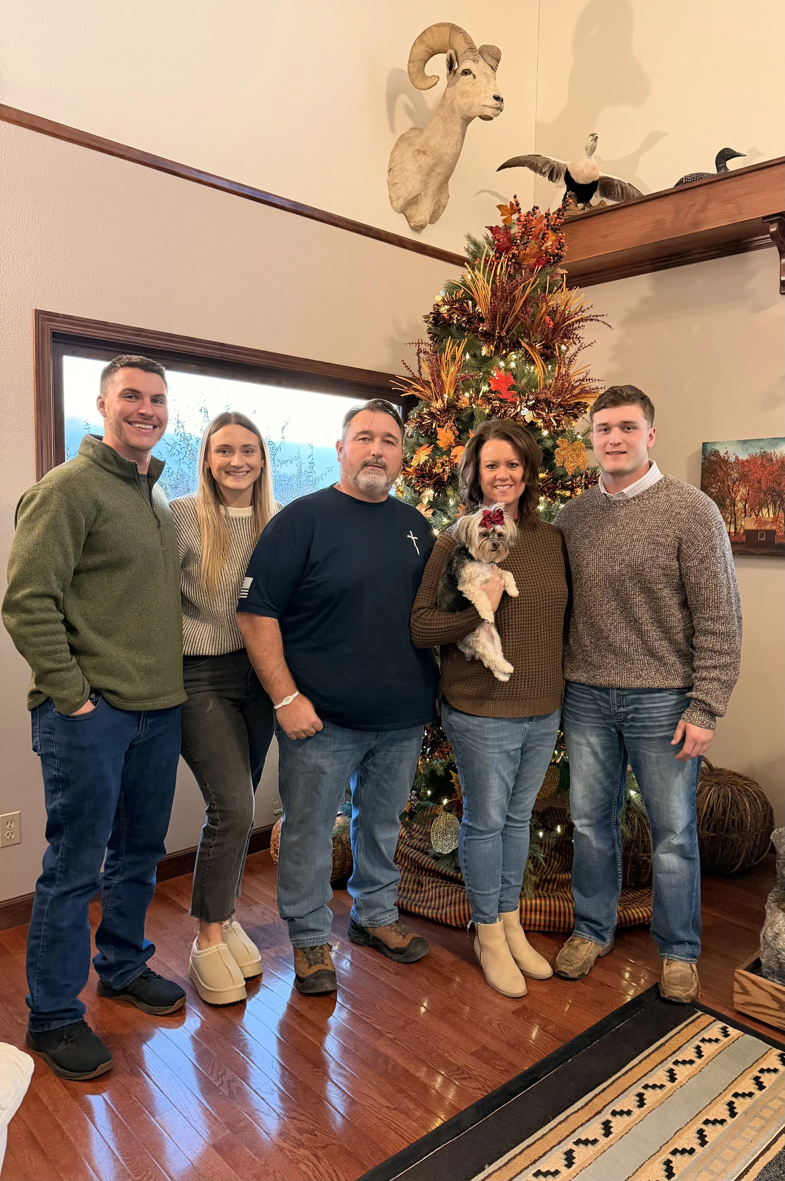 A family of five standing in front of a decorated Christmas tree with a small dog. They are indoors with wood flooring and a window with frost, and there are hanging animal mountings on the wall.