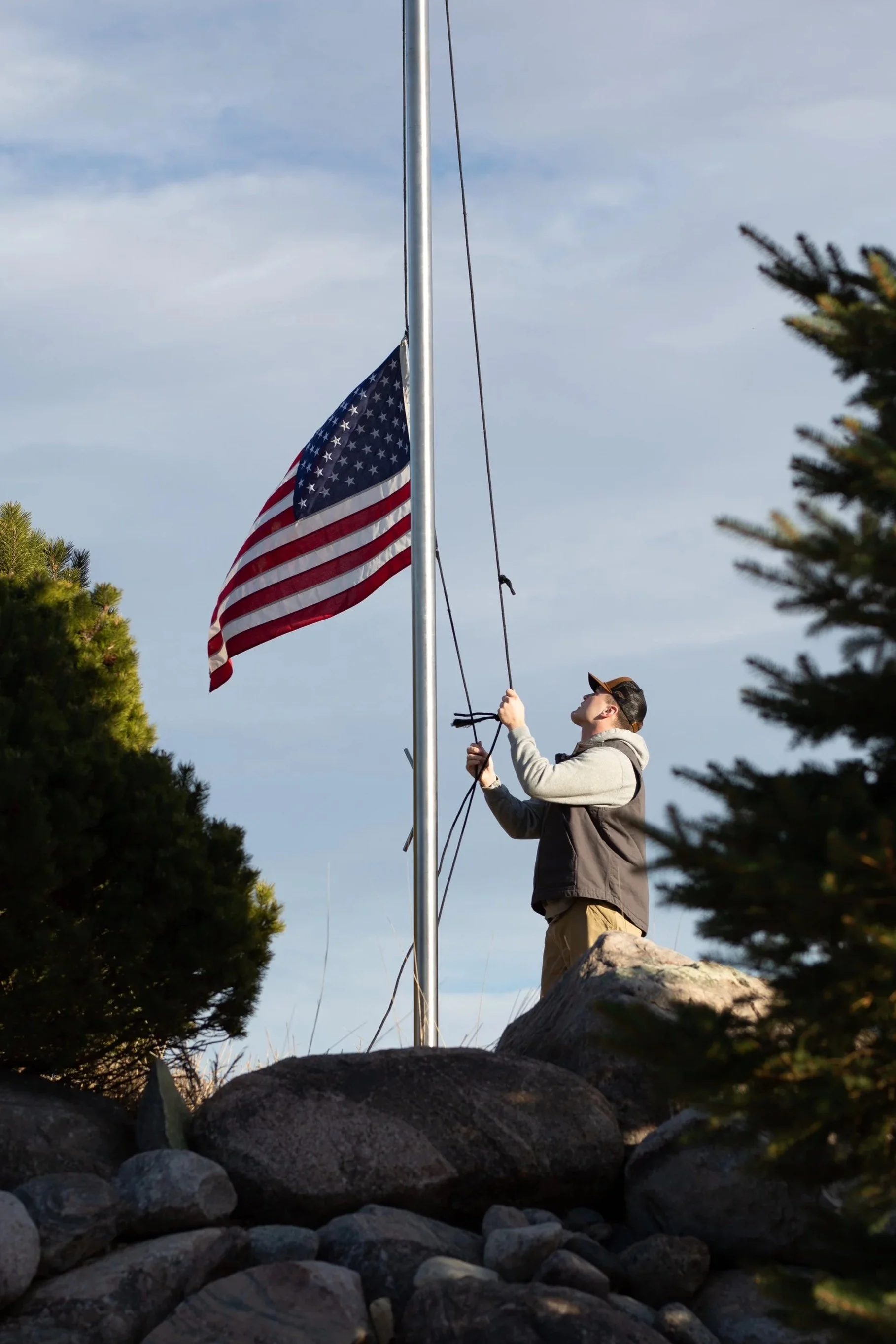 A person raising the American flag on a flagpole outdoors, surrounded by rocks and trees on a partly cloudy day.