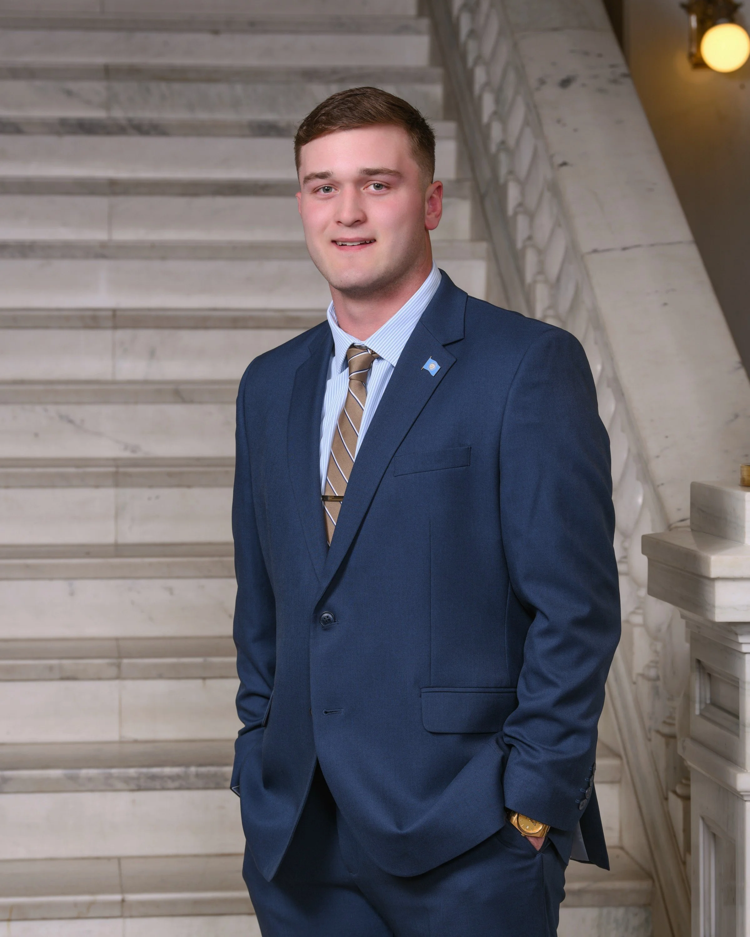 A young man in a navy blue suit, white and blue striped shirt, and a brown striped tie, standing on a marble staircase inside a building, with his right hand in his pocket and a gold watch on his left wrist.