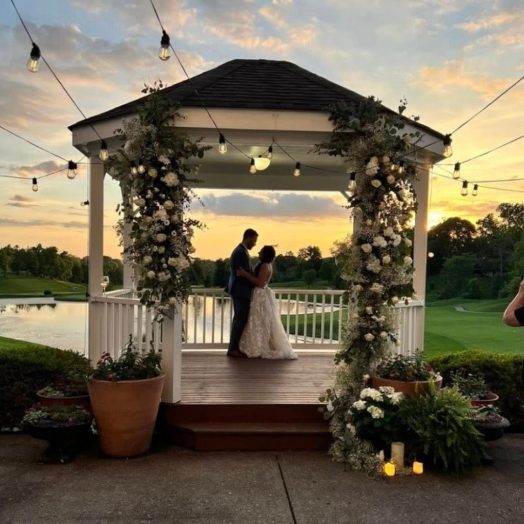 A couple getting married under a decorated gazebo at sunset, with string lights overhead and water in the background.