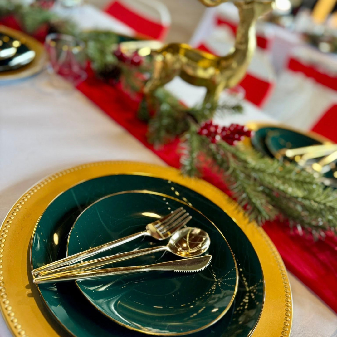 A festive holiday table setting with dark green plates and gold utensils on a yellow charger plate, decorated with Christmas greenery and red berries in the background.