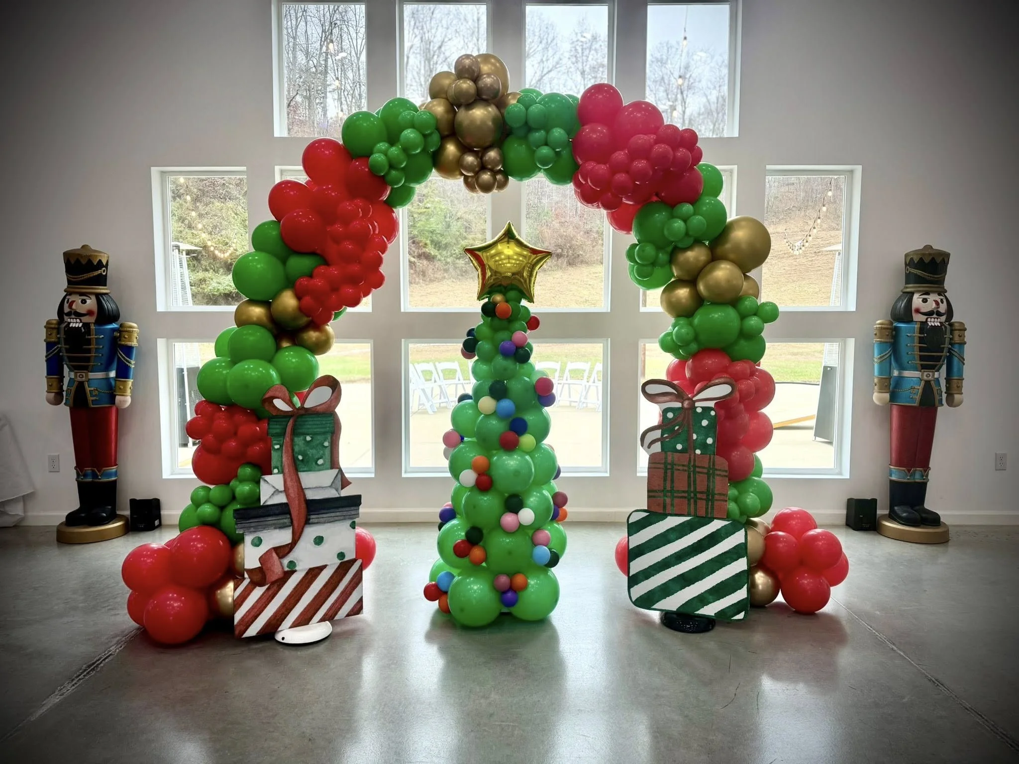 Colorful balloon arch with red, green, gold, and pink balloons, decorated with Christmas-themed cutouts of gifts and a star, set up in front of large windows, with nutcracker statues on either side.