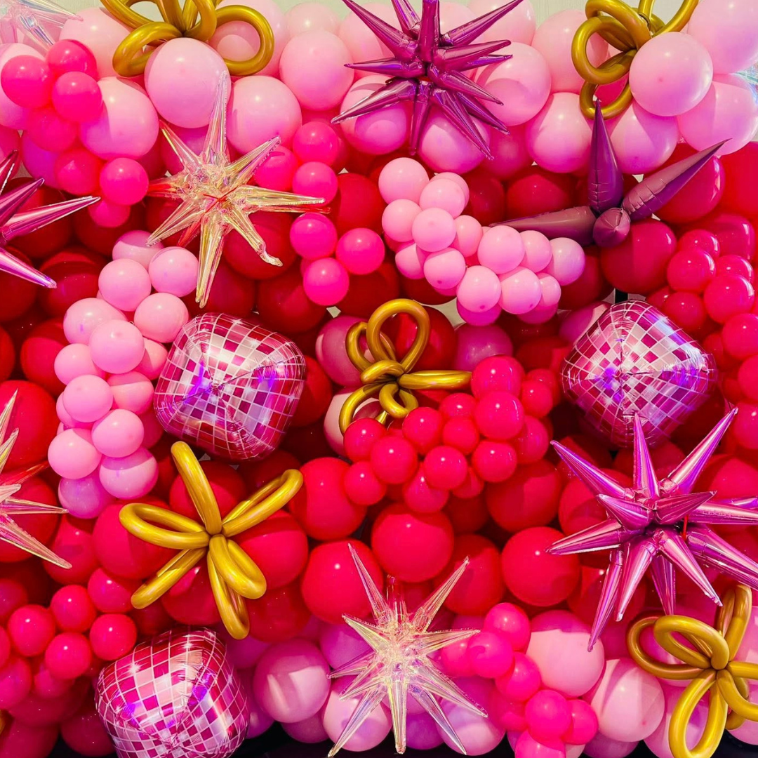 A vibrant arrangement of pink, purple, and gold balloons in various shapes, including star and heart shapes, creating a decorative and festive display.