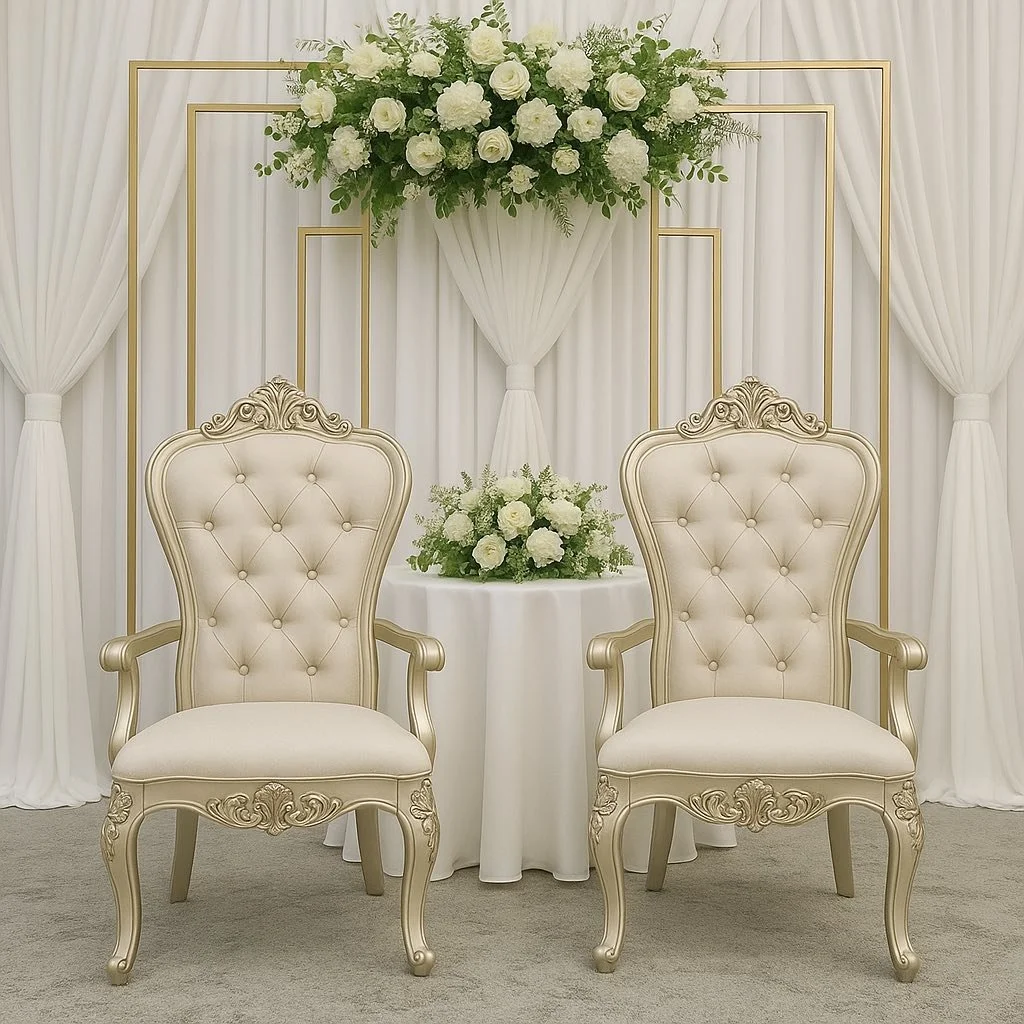 Elegant wedding ceremony setup with two ornate cream-colored chairs, a small round table with a floral arrangement, white curtains, and a floral display with white roses and greenery against a white backdrop.
