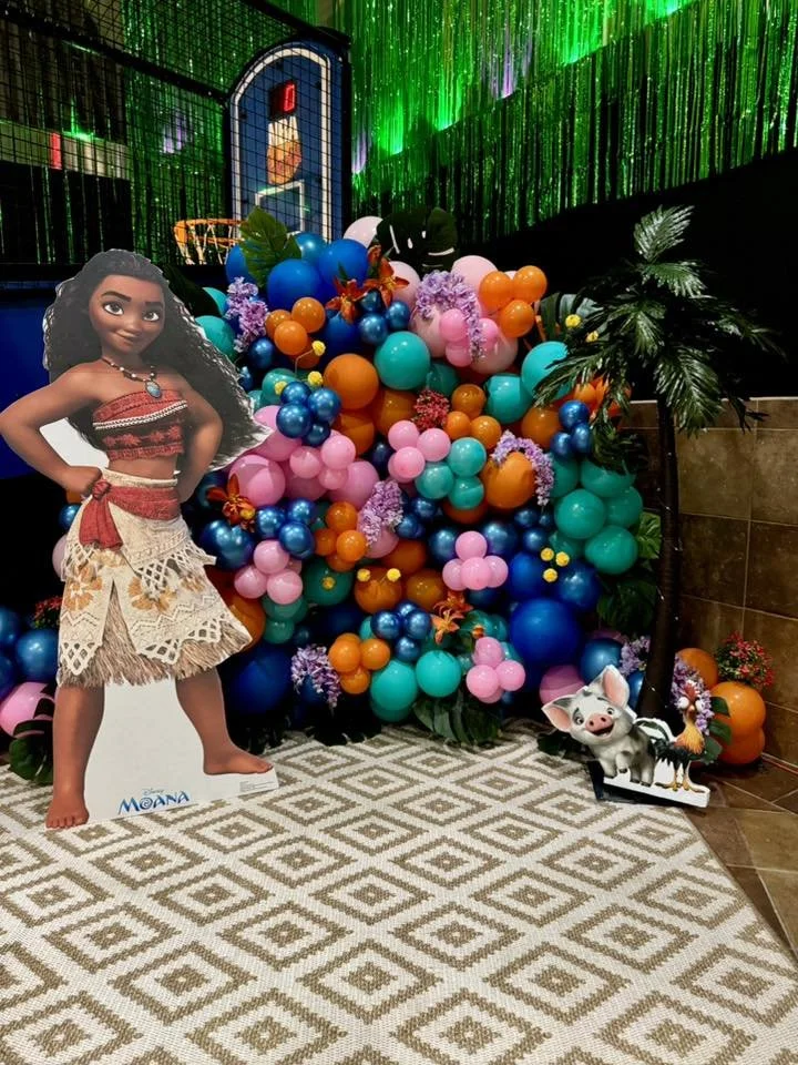 Colorful balloon display with pink, blue, orange, and purple balloons, and tropical leaf decorations. A cardboard cutout of Moana character on the left and a small pig and rooster character on the right. The background features green LED lights and a