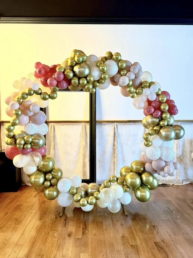 Large decorative balloon wreath with gold, white, pink, and speckled balloons, arranged in a circular shape, standing indoors on a wooden floor.