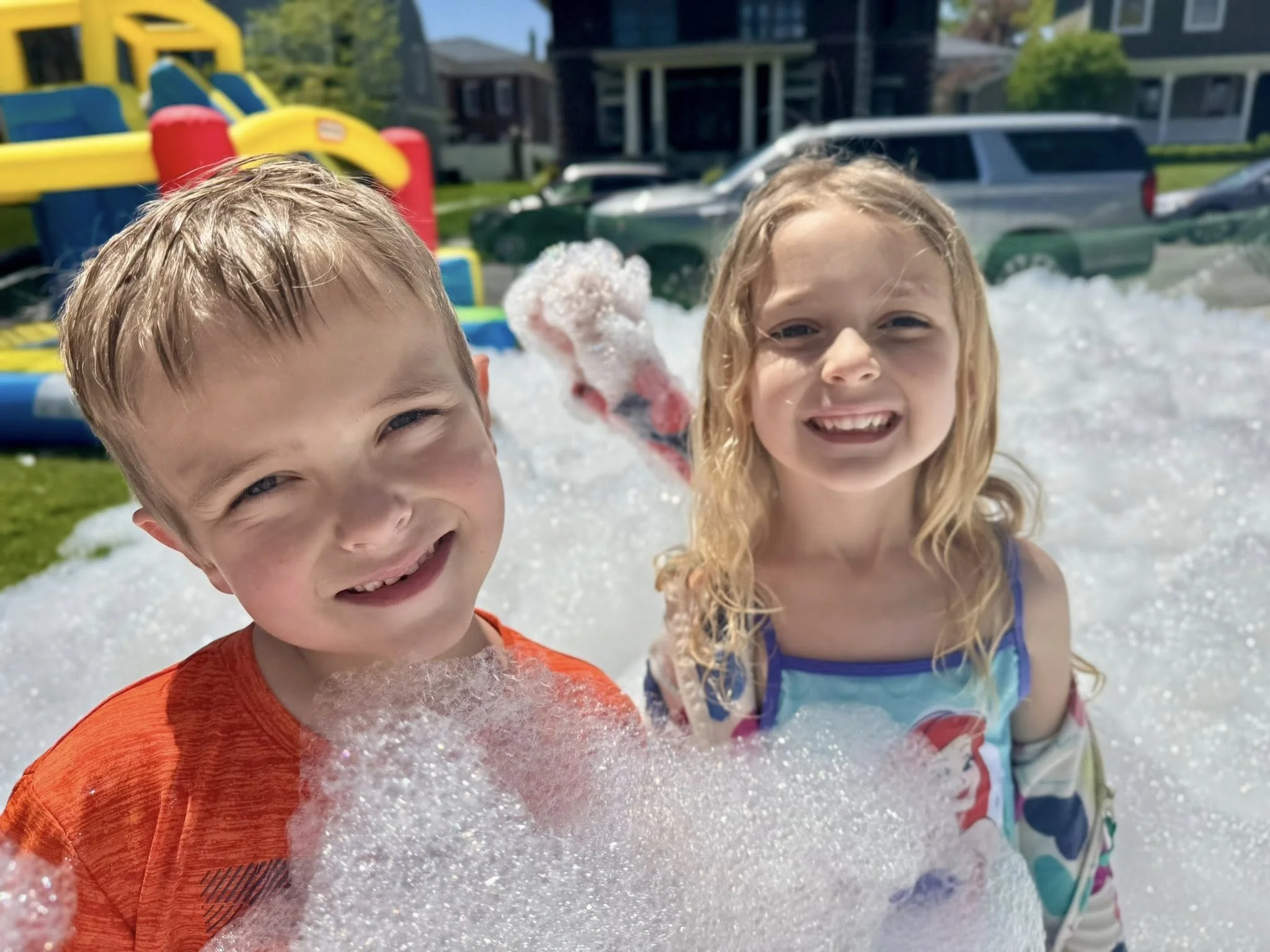 Two children, a boy in an orange shirt and a girl in a colorful dress, smiling and playing in a bubbly outdoor foam pool with a foam dispenser in the background.