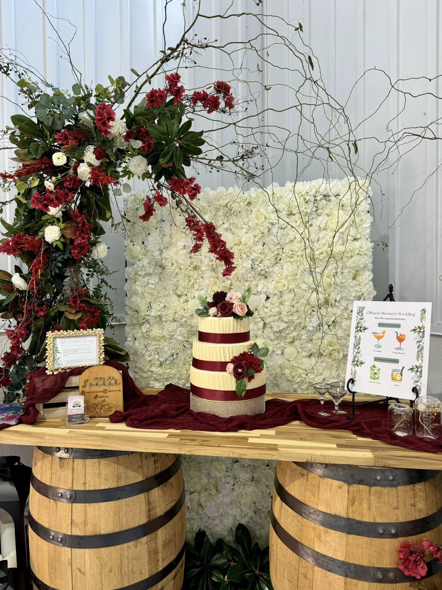 Wedding dessert table with a three-tiered cake decorated with red and pink flowers. The table is made of wood barrels with a red cloth, and there are floral arrangements, a drink menu, and decorative jars in front of a white floral backdrop.