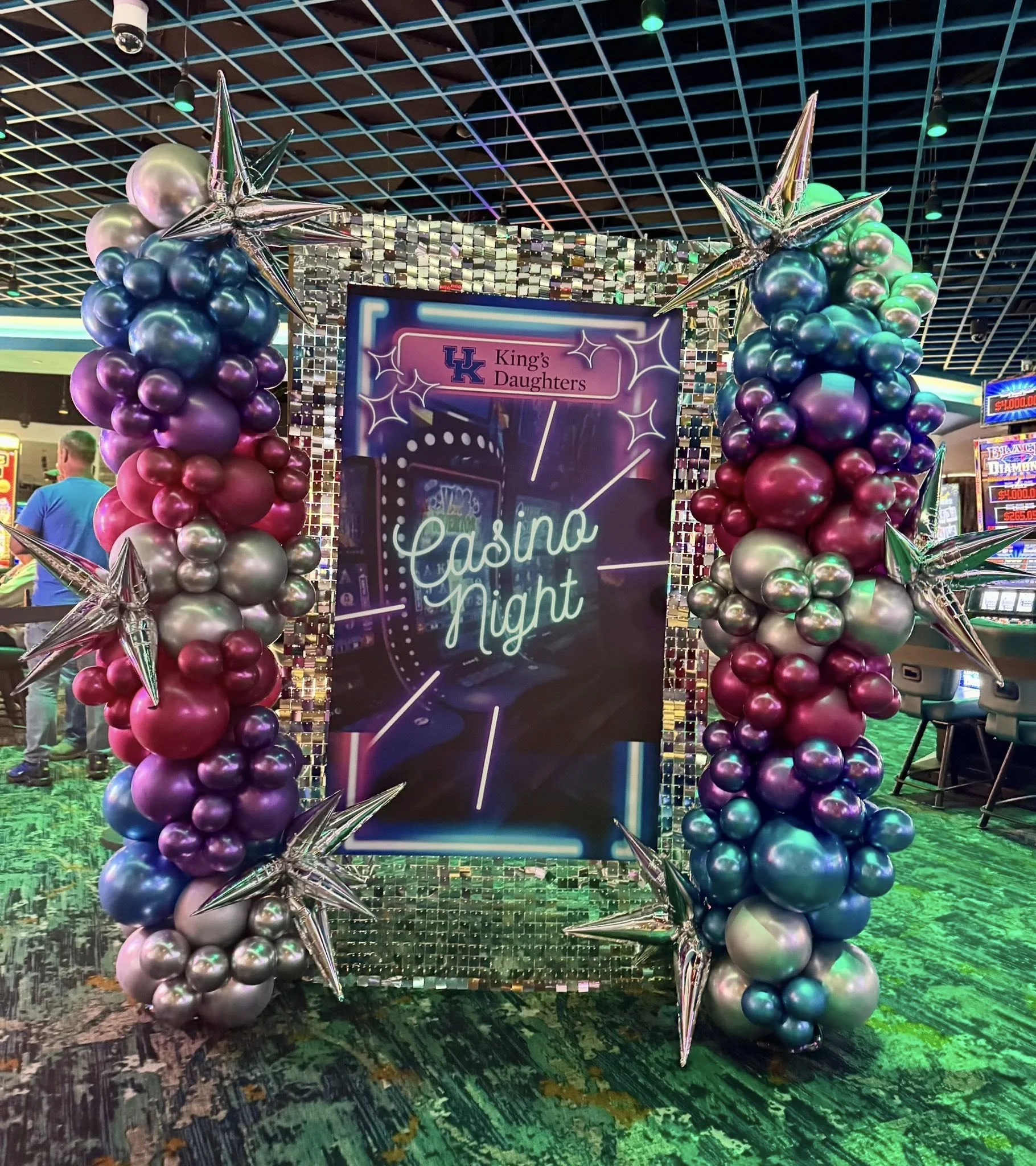 Decorative frame of purple, pink, silver balloons with silver star balloons, surrounding a neon-lit sign that reads 'King's Daughters Casino Night' in an indoor casino.