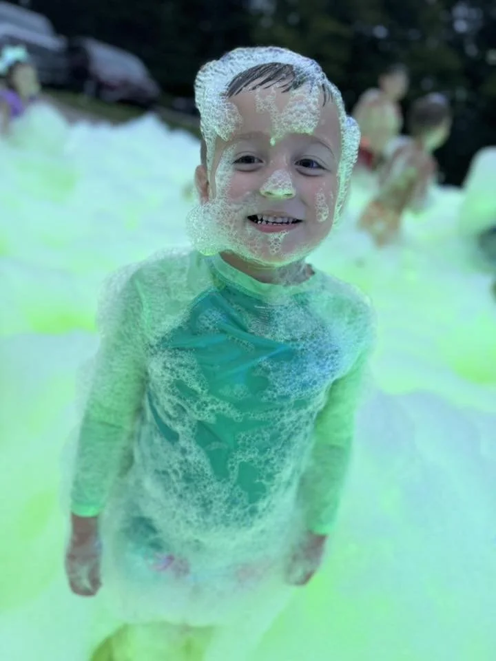 Young boy smiling in a foam-filled water park with glowing green water.