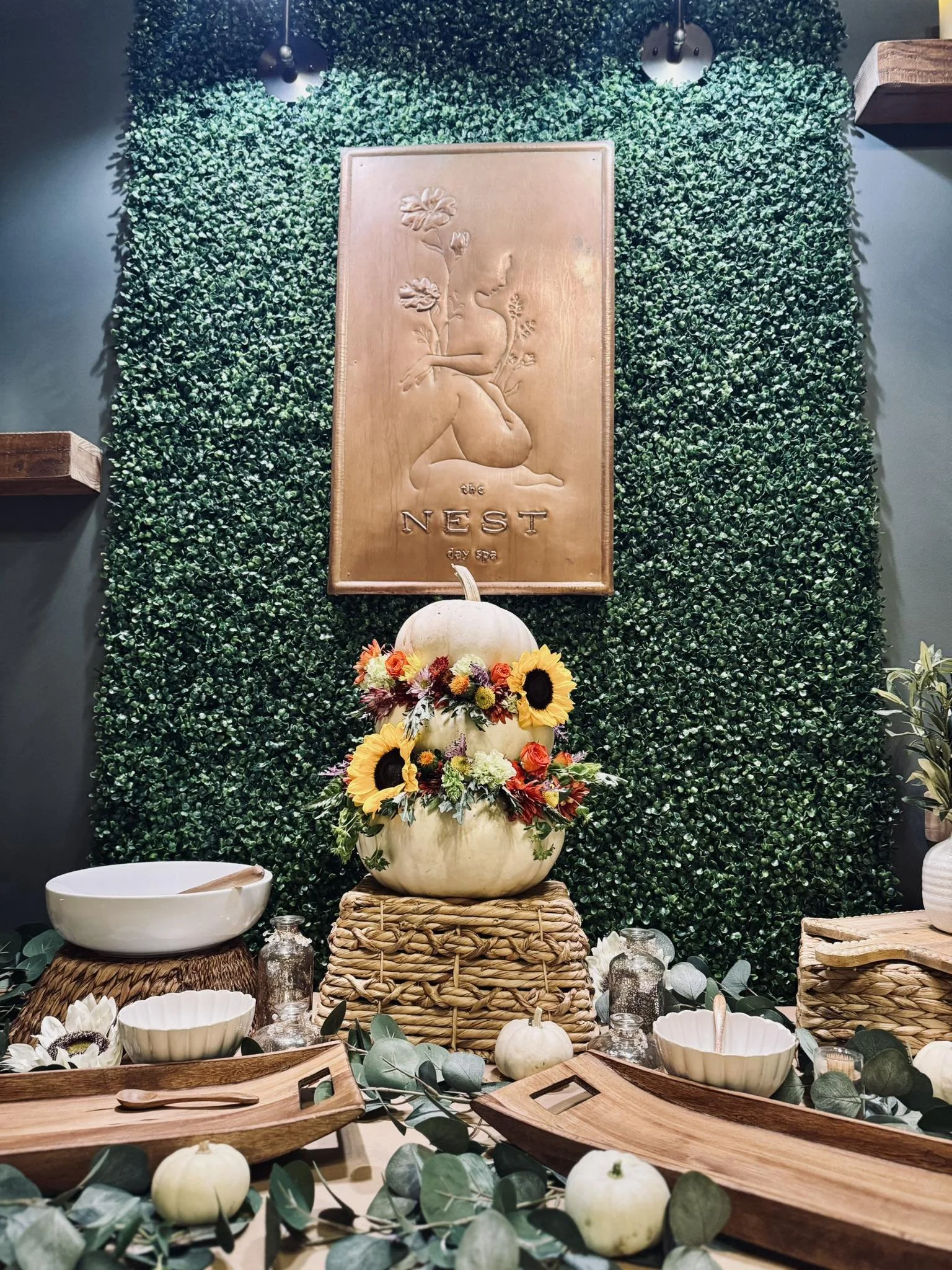 Decorative fall display at a spa named The Nest, featuring a large white pumpkin with sunflower and flower arrangements on top of a wicker platform, surrounded by smaller pumpkins, skewered bowls, eucalyptus leaves, and small jars, with a green leafy