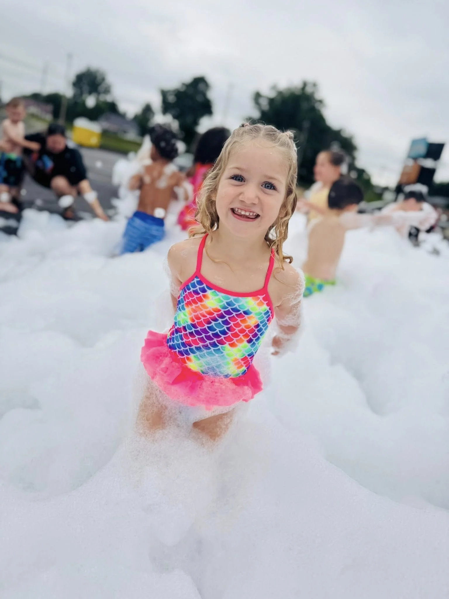 Young girl in a colorful swimsuit smiling in foam at a foam party, other children and adults in the background.