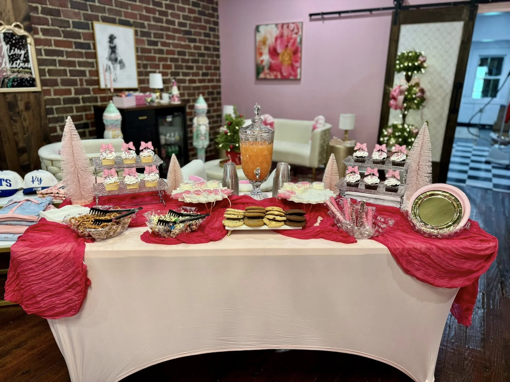 Party table decorated with pink and cream tablecloth, featuring cupcakes with pink bows, cookies, and a large orange drink dispenser, set in a room with pink and brick walls, decorated with pink roses and framed art.