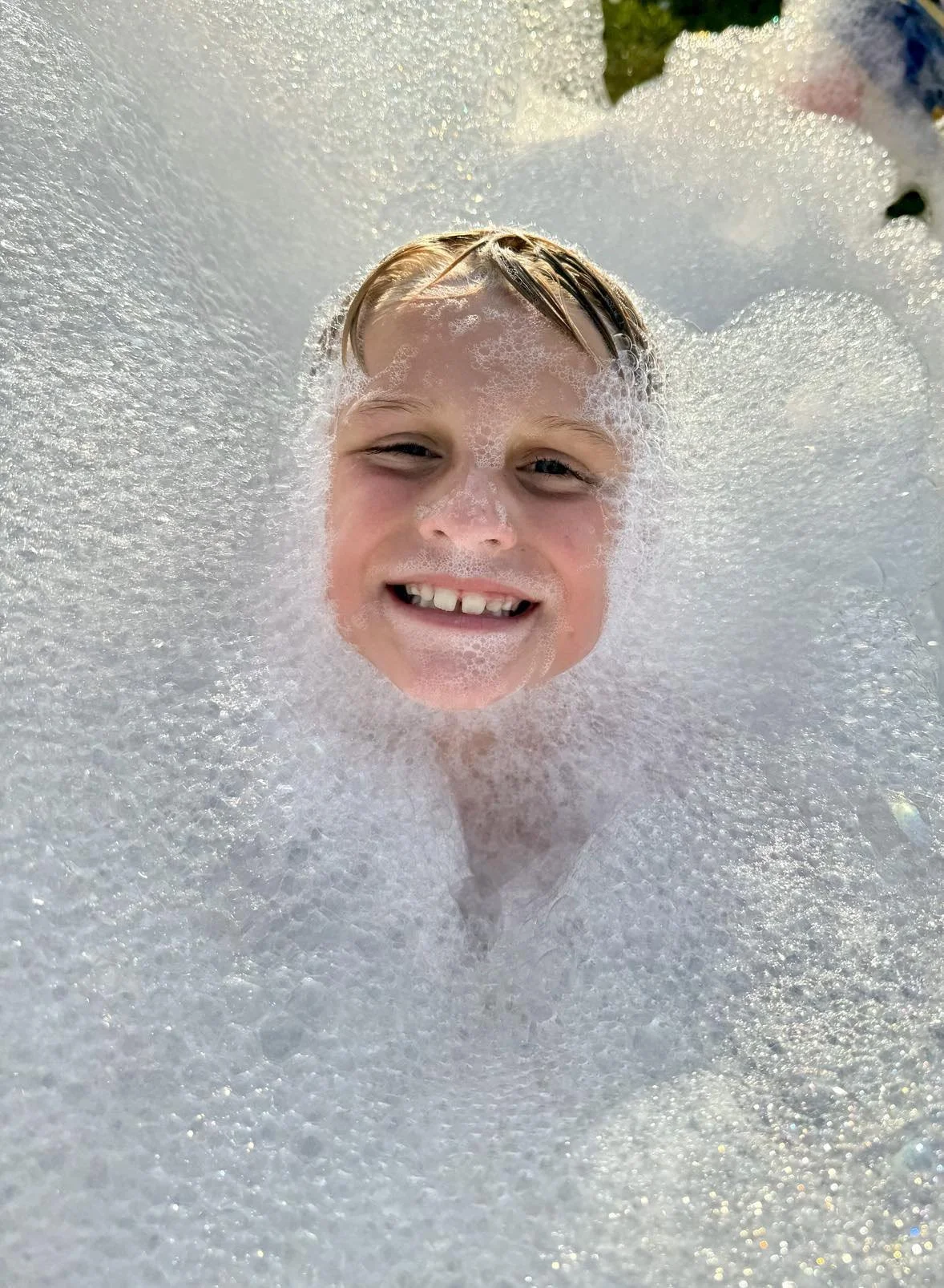 A smiling young boy with wet hair surrounded by white soap bubbles in a bubble bath.