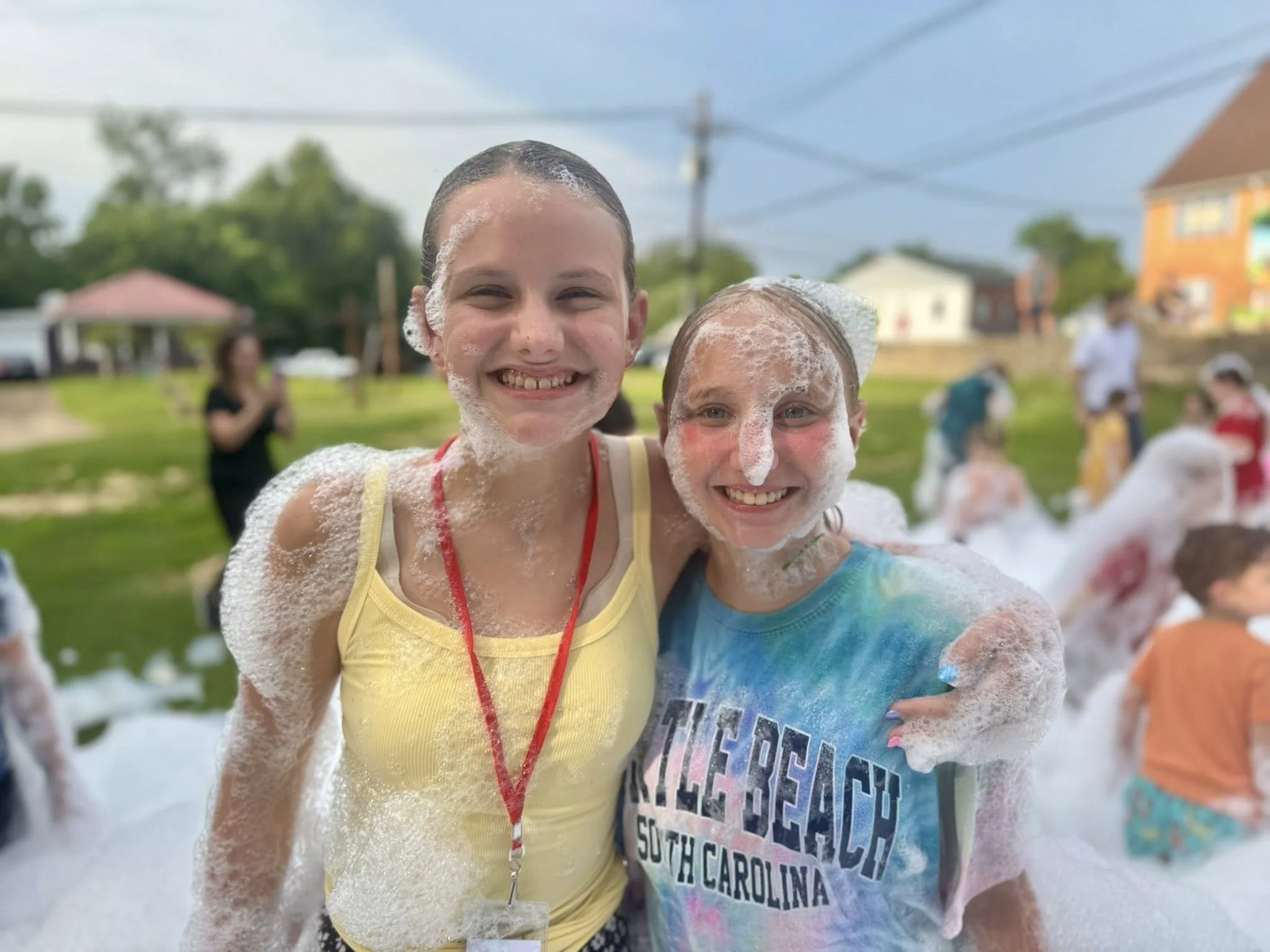 Two young girls smiling and covered in bubbles at an outdoor foam party. One girl is wearing a yellow tank top and the other a tie-dye shirt with text that says "MILE BEACH SOUTH CAROLINA." In the background, people are enjoying the event on a grassy