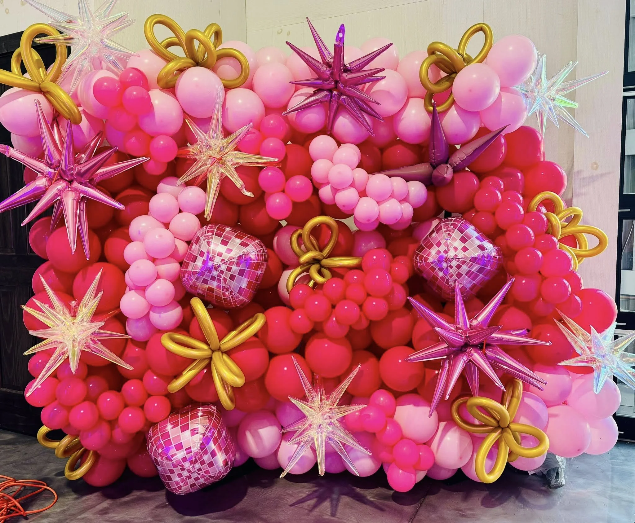 A vibrant balloon arrangement featuring various shades of pink and gold balloons, decorated with star-shaped metallic balloons in pink, silver, and purple, along with disco ball patterned balloons, creating a colorful and festive display.