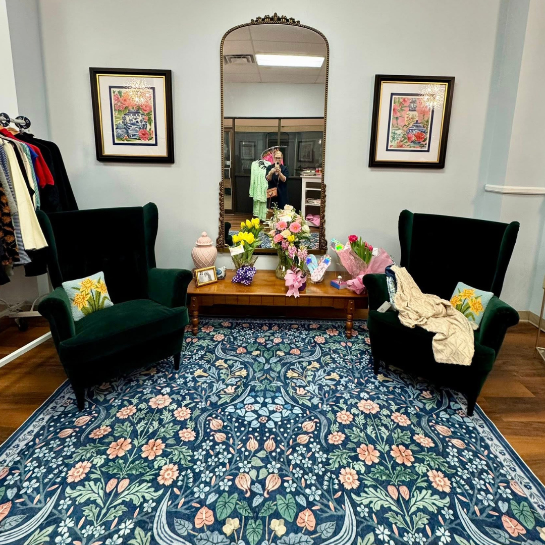 A cozy seating area with two dark green velvet armchairs with floral cushions, a wooden table with flower arrangements, and a large ornate mirror reflecting the photographer. The wall is decorated with two framed floral artwork.