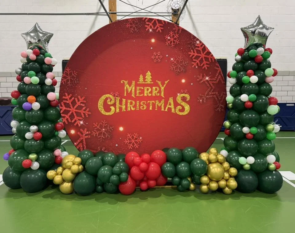 Christmas balloon display with two decorated Christmas trees made of balloons and a large red circular backdrop with 'Merry Christmas' written on it, featuring snowflake designs.