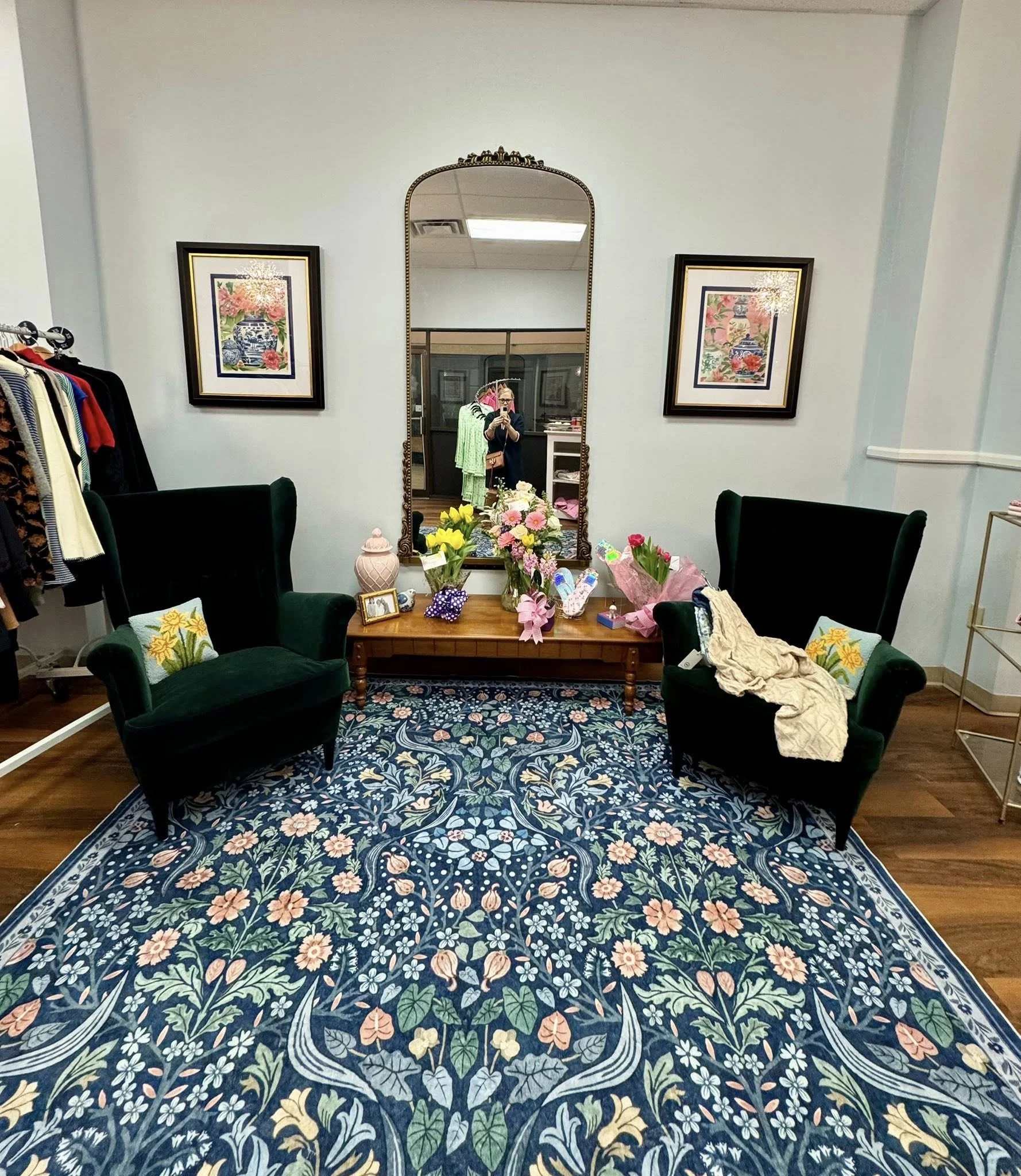 A cozy boutique fitting room with two dark green velvet armchairs, a floral area rug, and a wooden table with flowers and personal items. A large ornate mirror reflects a person taking a photo. There are framed floral prints on the light-colored wall