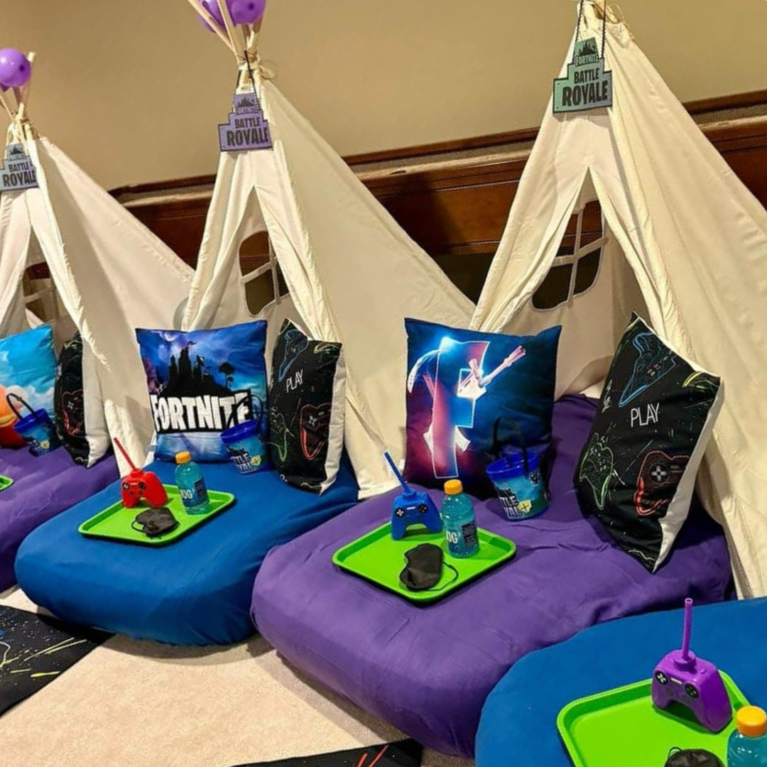 Three child-sized beds with Fortnite-themed pillows and tent-like canopies. Each bed has a colorful gaming controller, a bottle of water, and a small black item on a green tray, set against a beige wall.