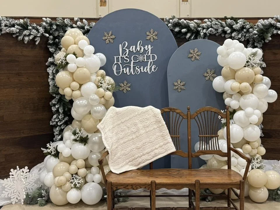 Baby shower backdrop with a blue rounded panel reading 'Baby It's Cold Outside,' surrounded by snowflake decorations and a balloon arch of white and beige balloons with frosted details. There is a wooden bench with a white knitted blanket in front of
