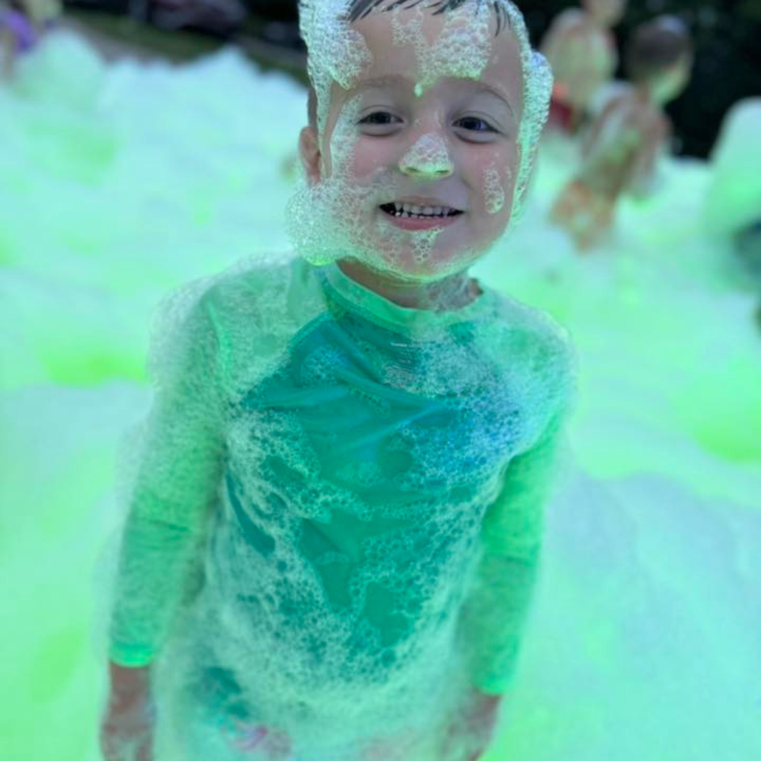 A smiling young boy covered in green and blue soap bubbles, surrounded by more children in the background, in a colorful foam party.