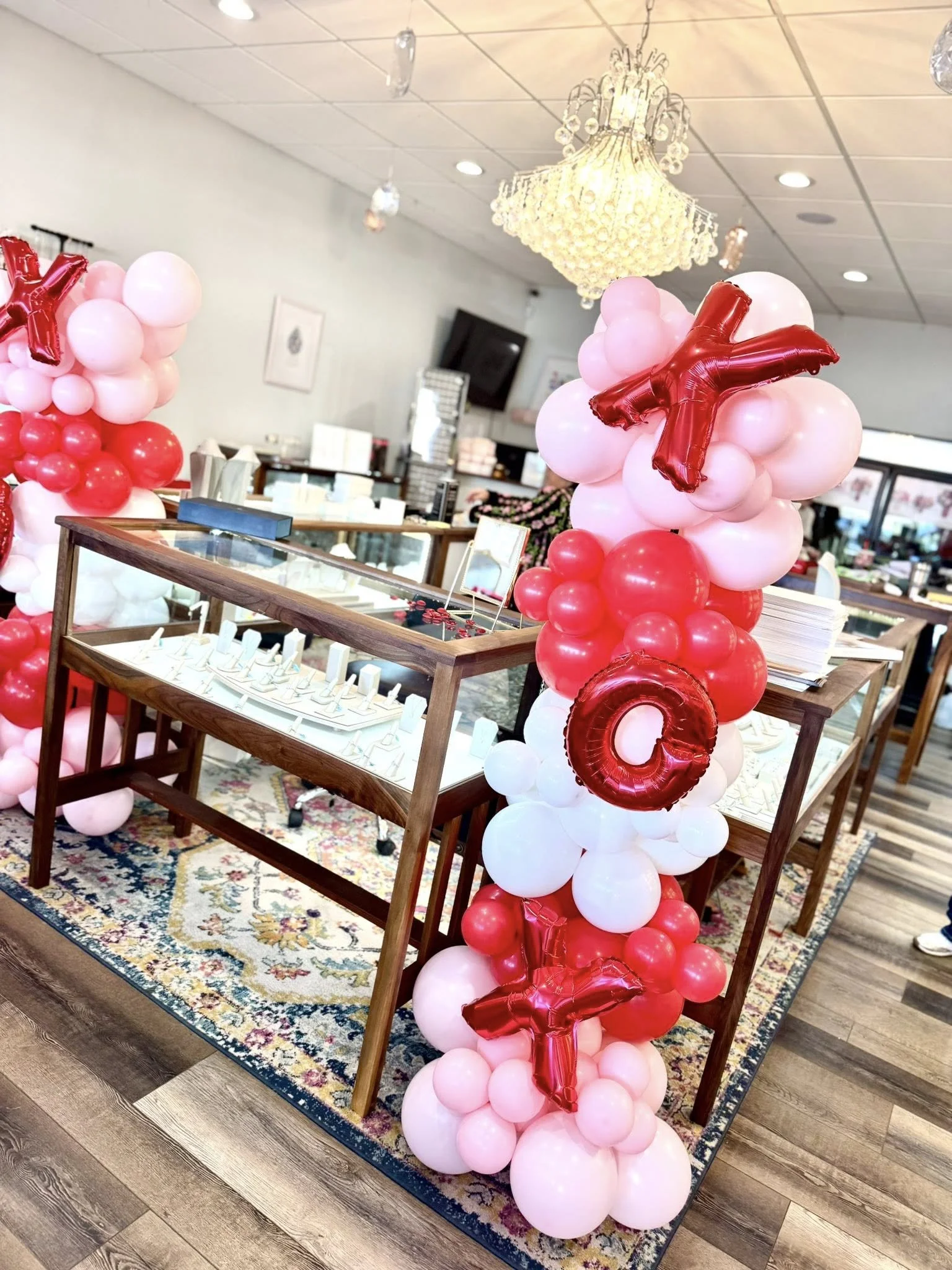 Decorative balloon columns with pink, white, and red balloons, featuring red foil balloons spelling out "Y" and "Q" placed around a wooden display table inside a decorated room.