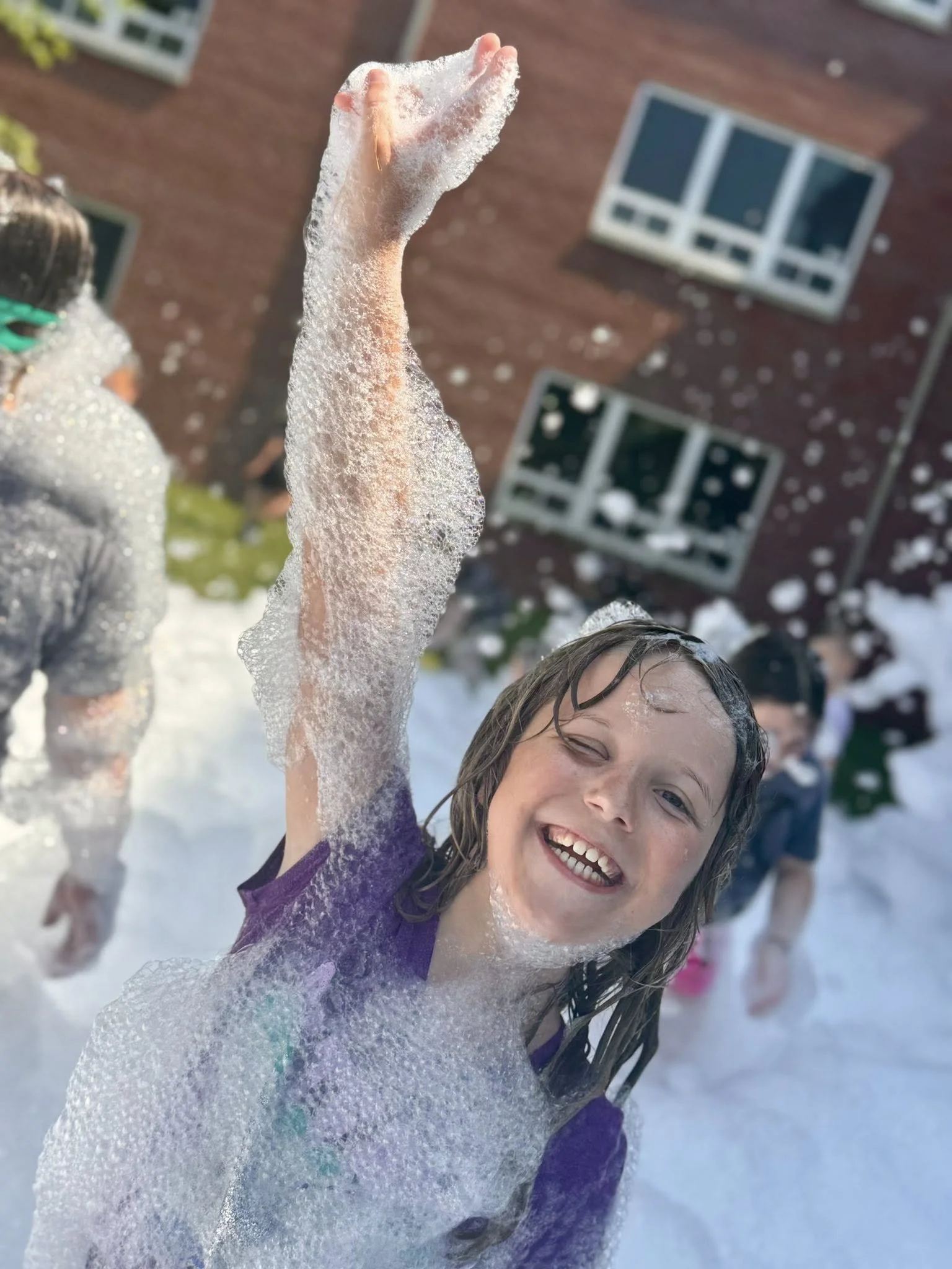 Smiling girl covered in soap bubbles playing in the foam outside with other children in the background.