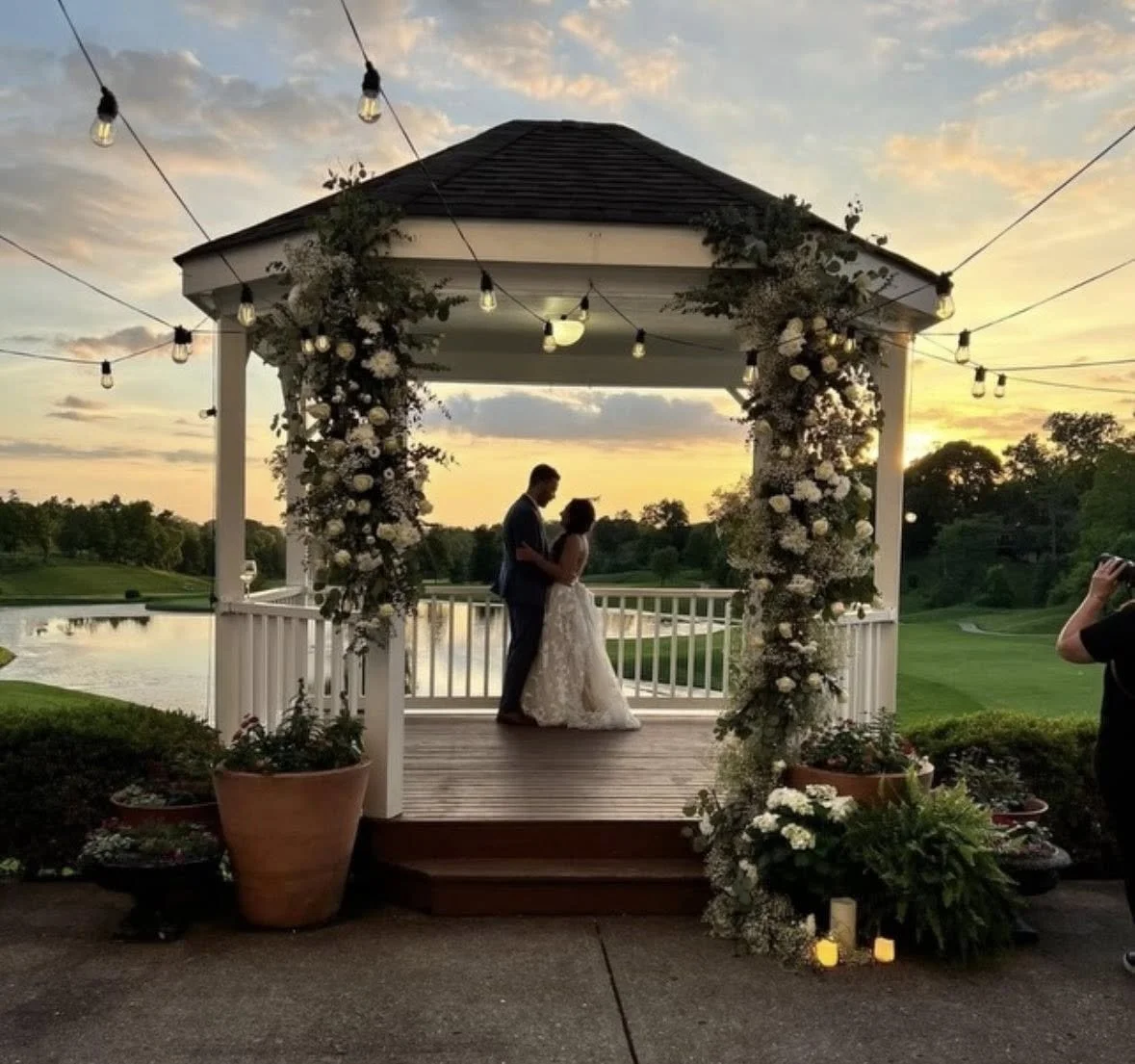 Couple getting married on a small gazebo by a lake at sunset, decorated with flowers and string lights, with a photographer capturing the moment.