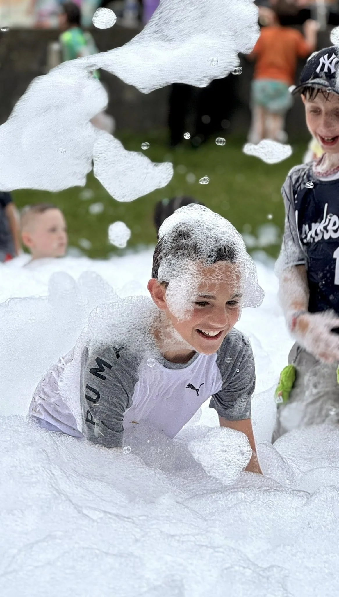 Children playing and laughing in a foam party outdoors, surrounded by bubbles and foam.