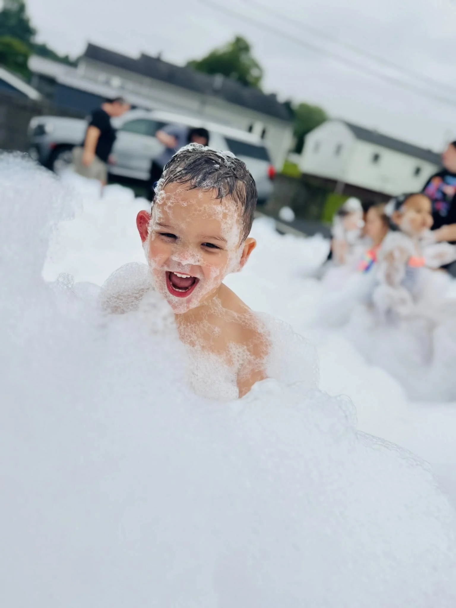 A young boy with foam on his face and head smiling and playing in a large foam bubble bath outdoors, with other children and adults in the background.