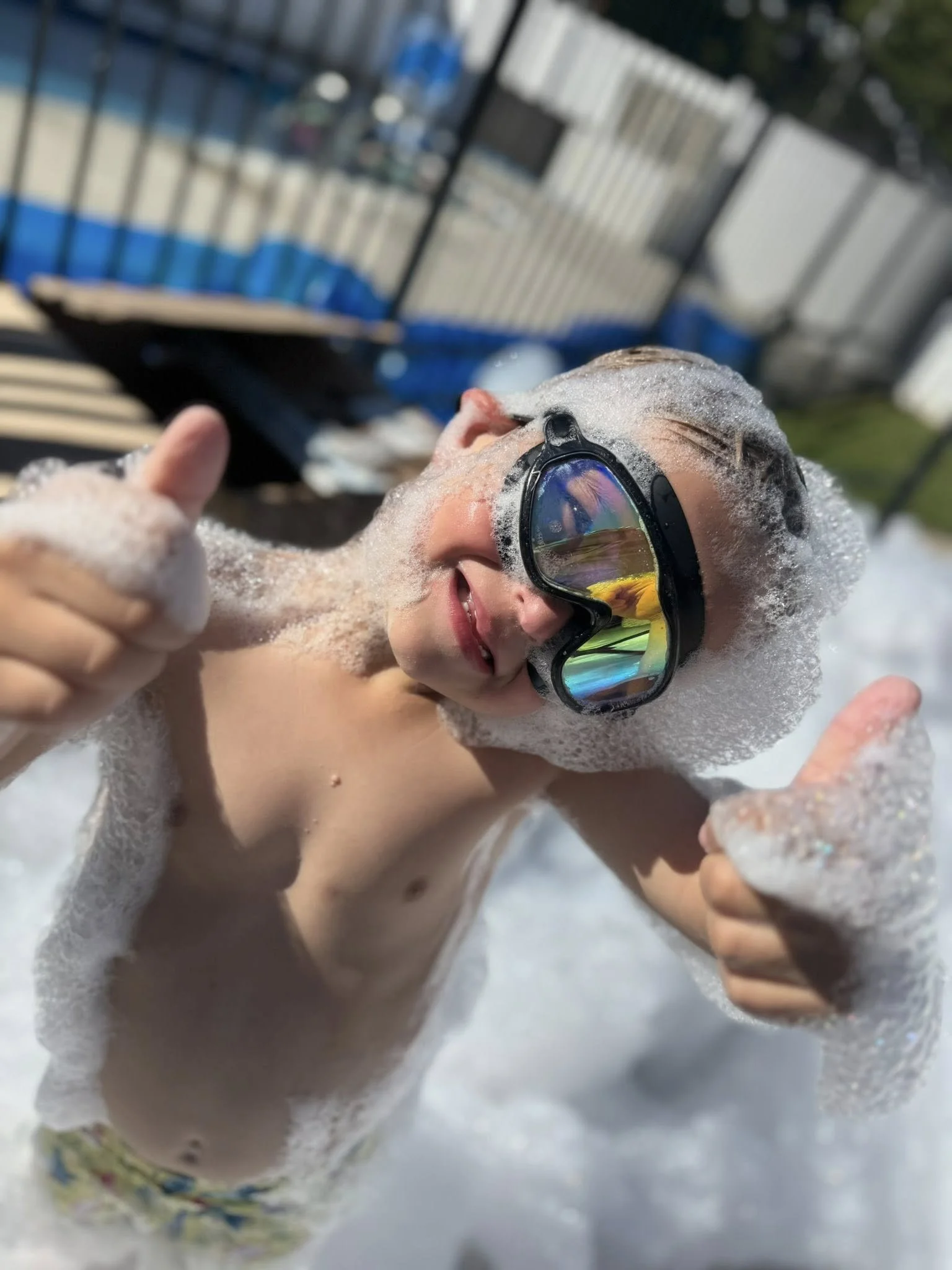 Child in foam and goggles giving thumbs up at a pool or splash pad during daytime.