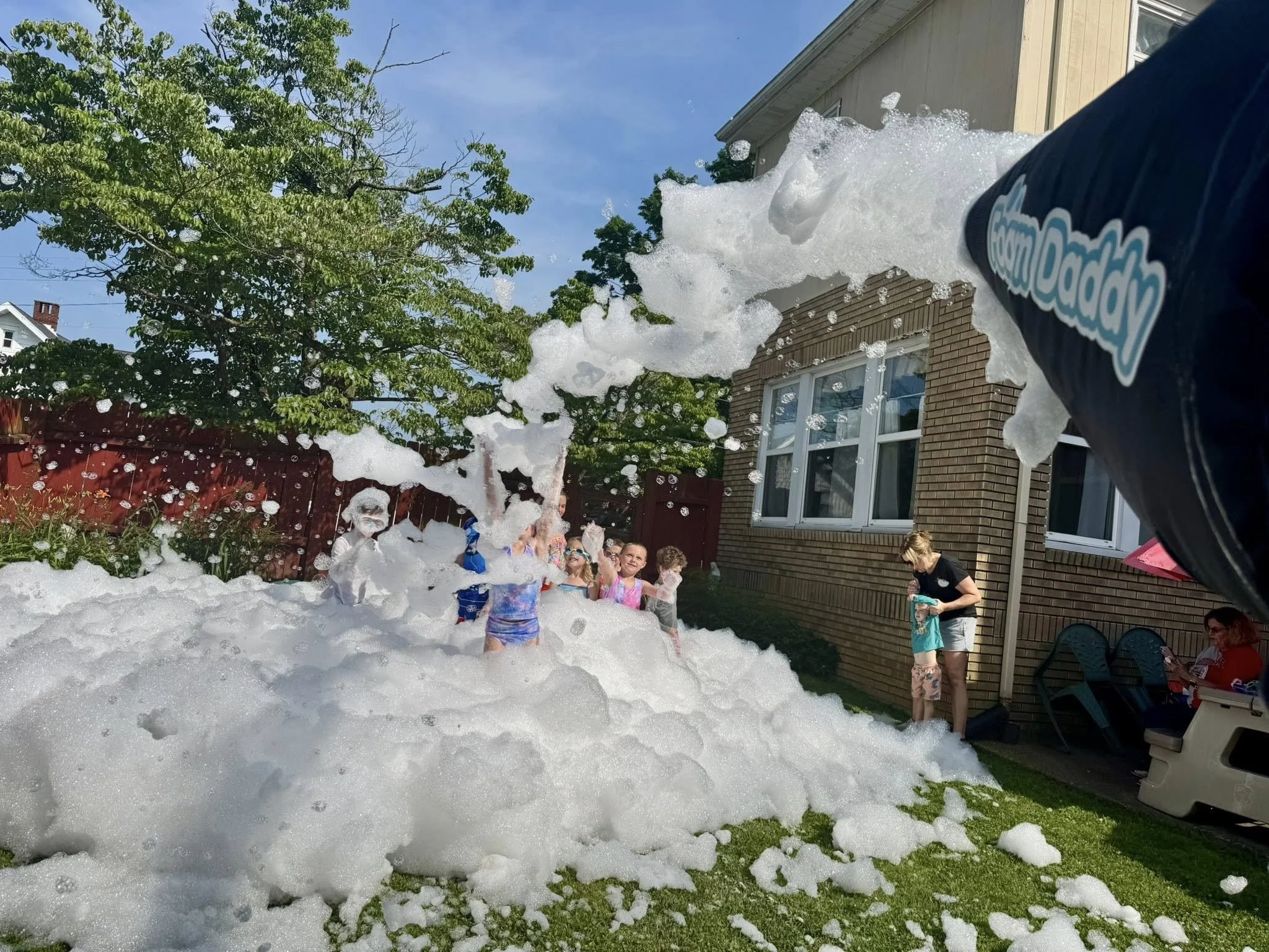Children and adults enjoying a foam party in a backyard with a brick house, green grass, and trees on a sunny day.
