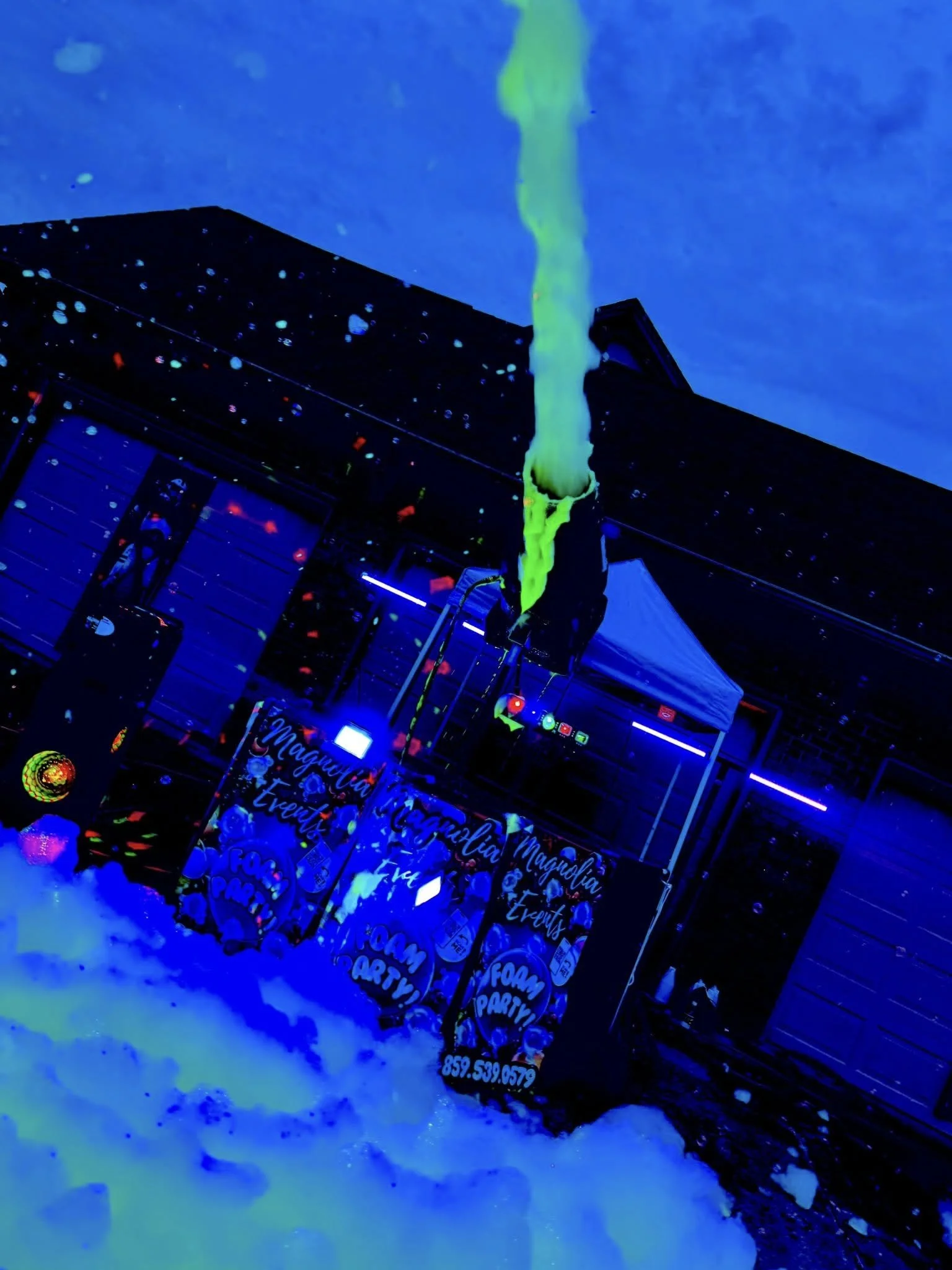 Outdoor foam party setup with foam falling from a machine, foam on the ground, illuminated by black lights, with a sign reading "Magnolia Events Foam Party!".