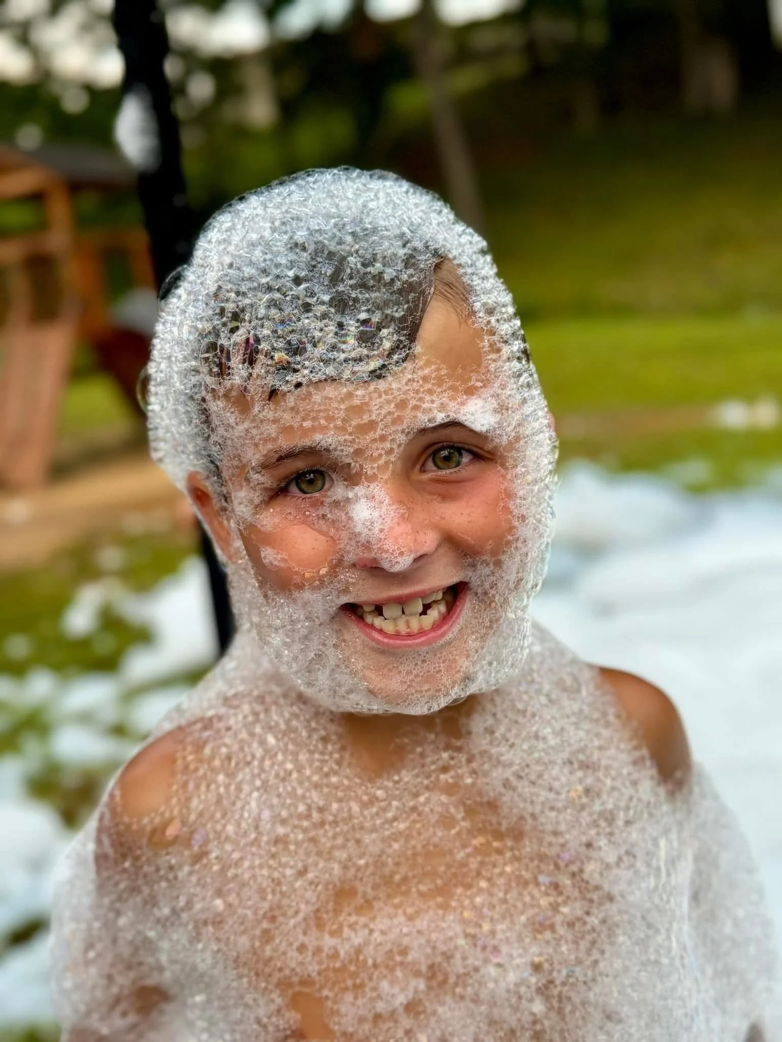 A young boy with soap bubbles on his face and body, smiling in an outdoor setting with blurred trees and a wooden structure in the background.