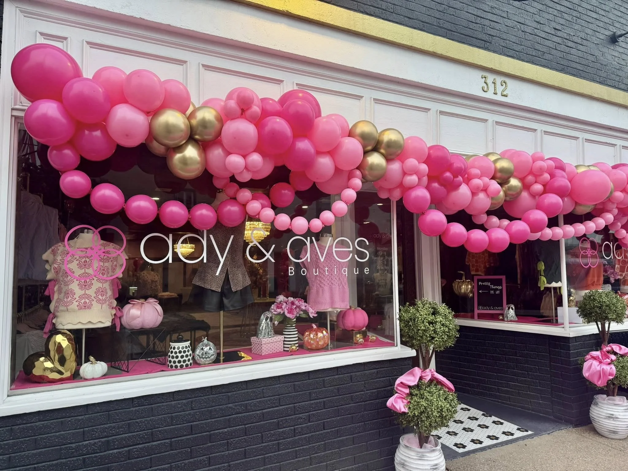 Storefront of a boutique named 'ady & aves' decorated with pink and gold balloons and potted plants with pink bows in front.