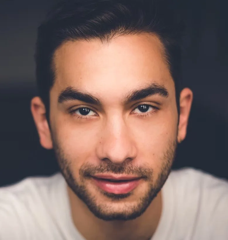 Close-up of a young man with dark hair, light beard, and blue eyes, smiling slightly, wearing a white shirt.