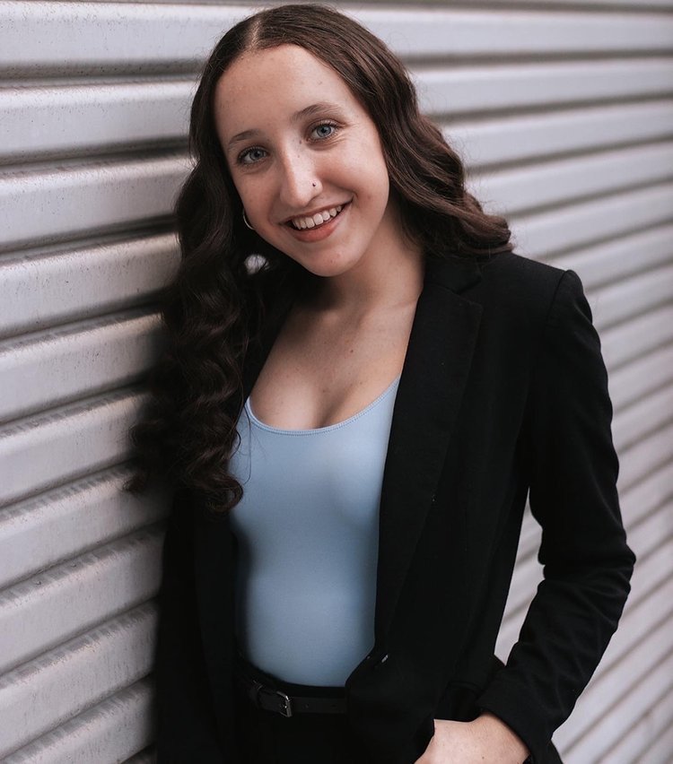 A young woman with long brown wavy hair, blue eyes, and a nose piercing, smiling while leaning against a metal wall. She is wearing a black blazer over a light blue top.