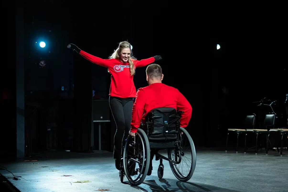 A woman in a red shirt and black pants performs a dance move for a man in a wheelchair on a stage with black background and stage lights.
