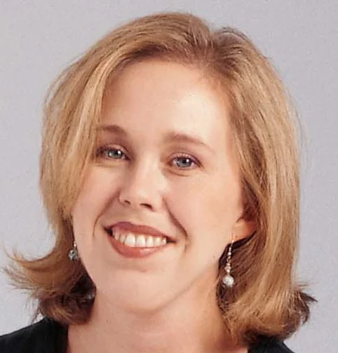 A woman with shoulder-length blonde hair smiling, wearing earrings, against a neutral background.