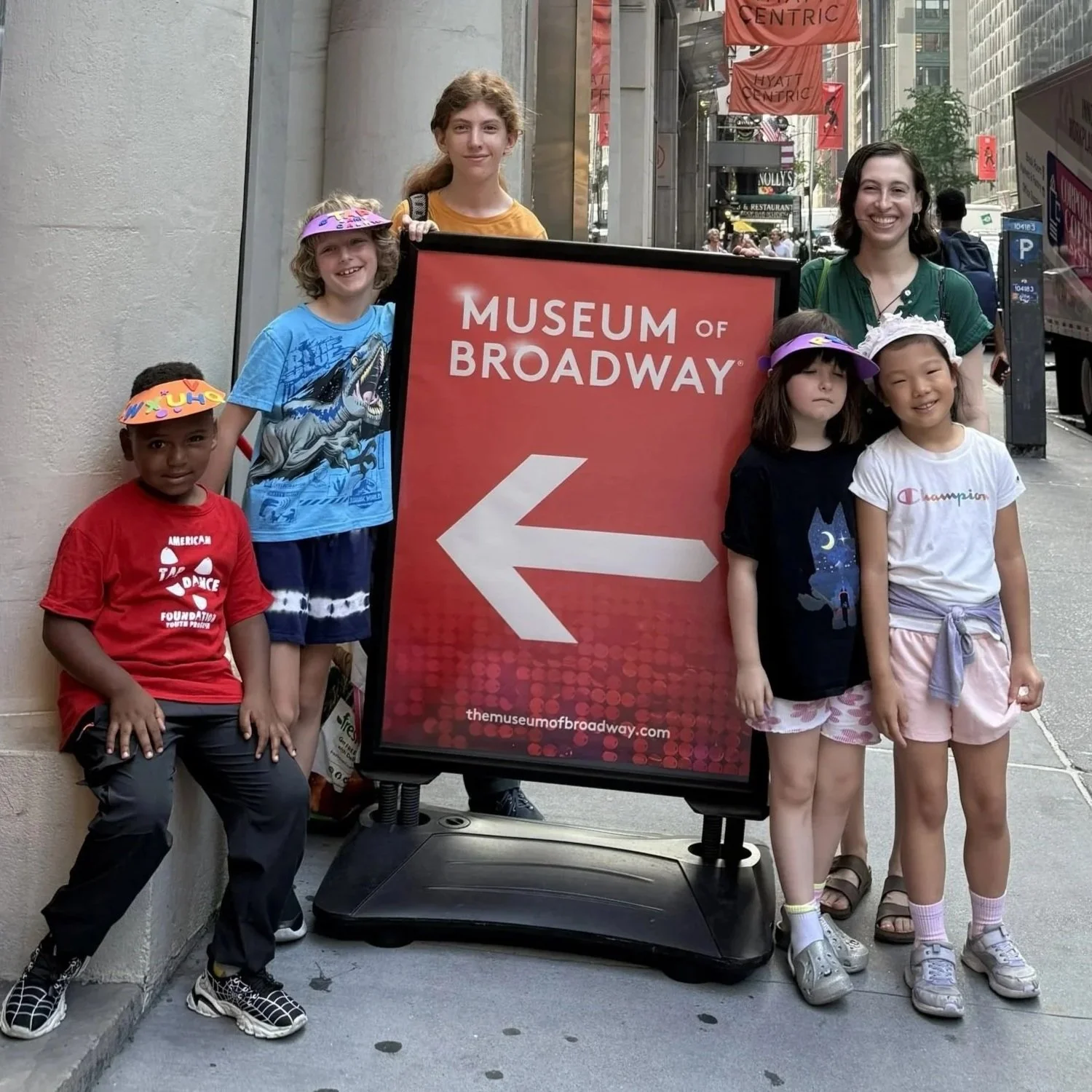 A group of children and a woman are standing on a city sidewalk next to a sign for the Museum of Broadway, which has an arrow pointing to the left. The children are smiling and wearing casual summer clothes and hats. The woman is smiling and wearing a green top.