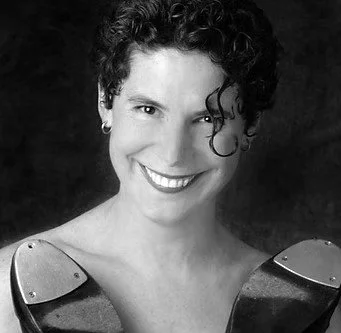 Black and white portrait of a woman with short, curly hair, smiling, wearing a top with metallic shoulder accents.