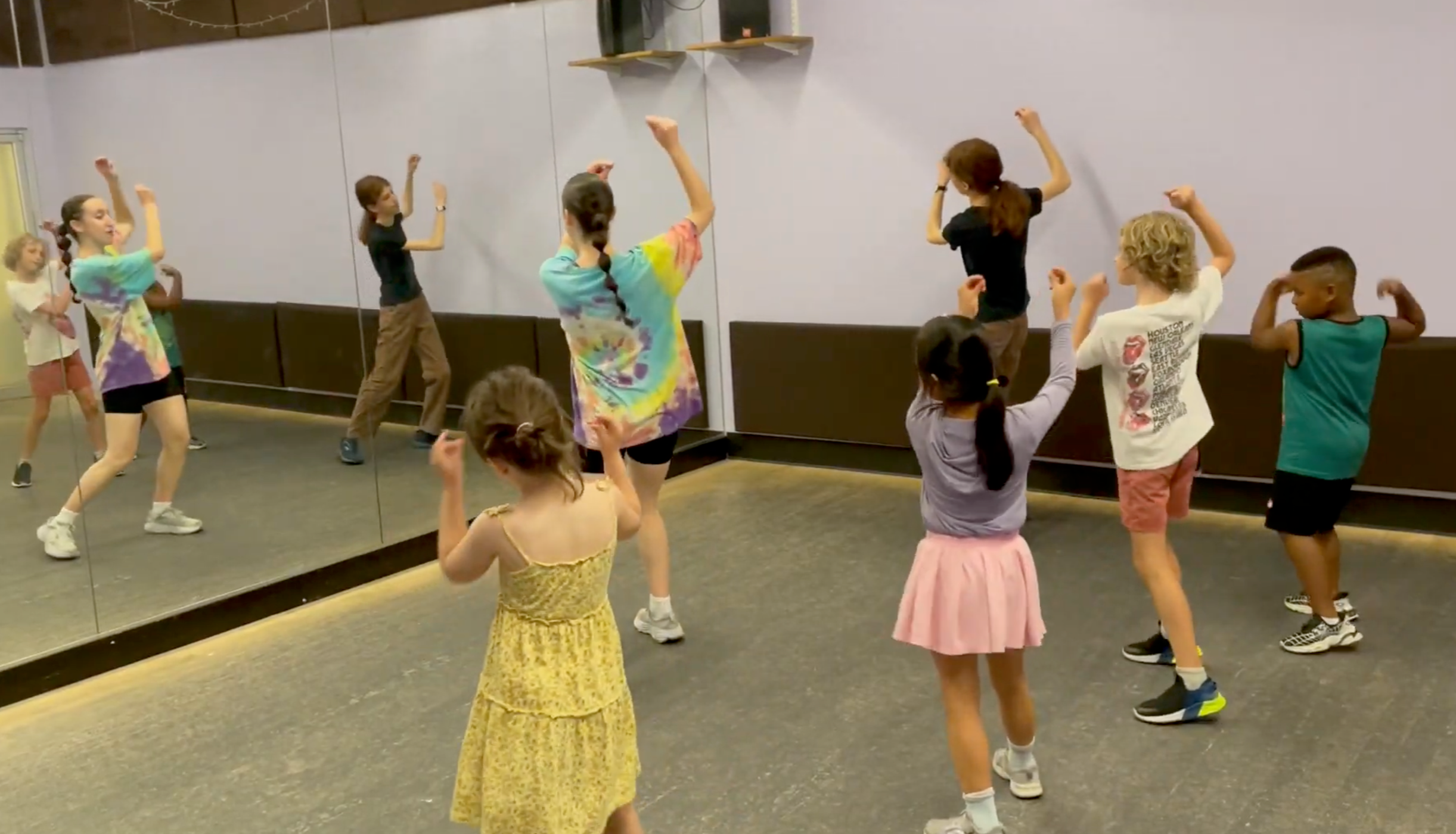 Children participating in a dance class, mimicking dance moves in front of a mirror in a studio.