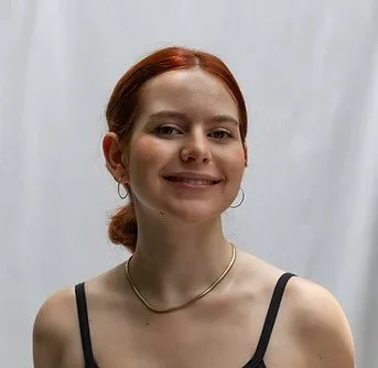 A young woman with red hair styled in a low bun, wearing a black tank top, a thin gold necklace, and hoop earrings, smiling at the camera against a plain light-colored background.