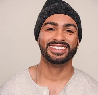 A man with a beard and mustache wearing a black beanie and a light gray shirt, smiling at the camera.